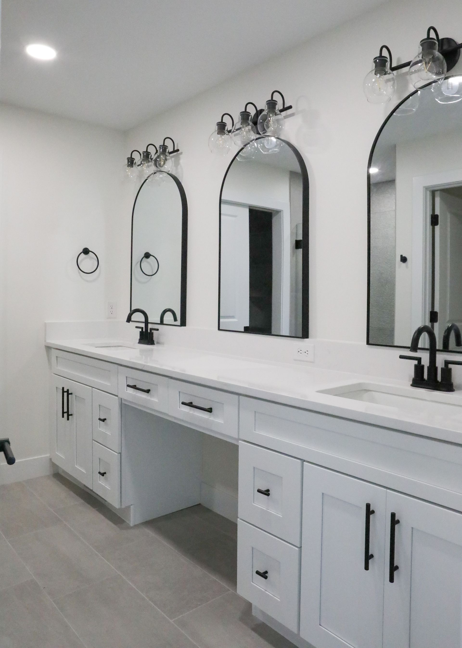 White bathroom vanity with three arched mirrors, black sconces, and gray tile floor