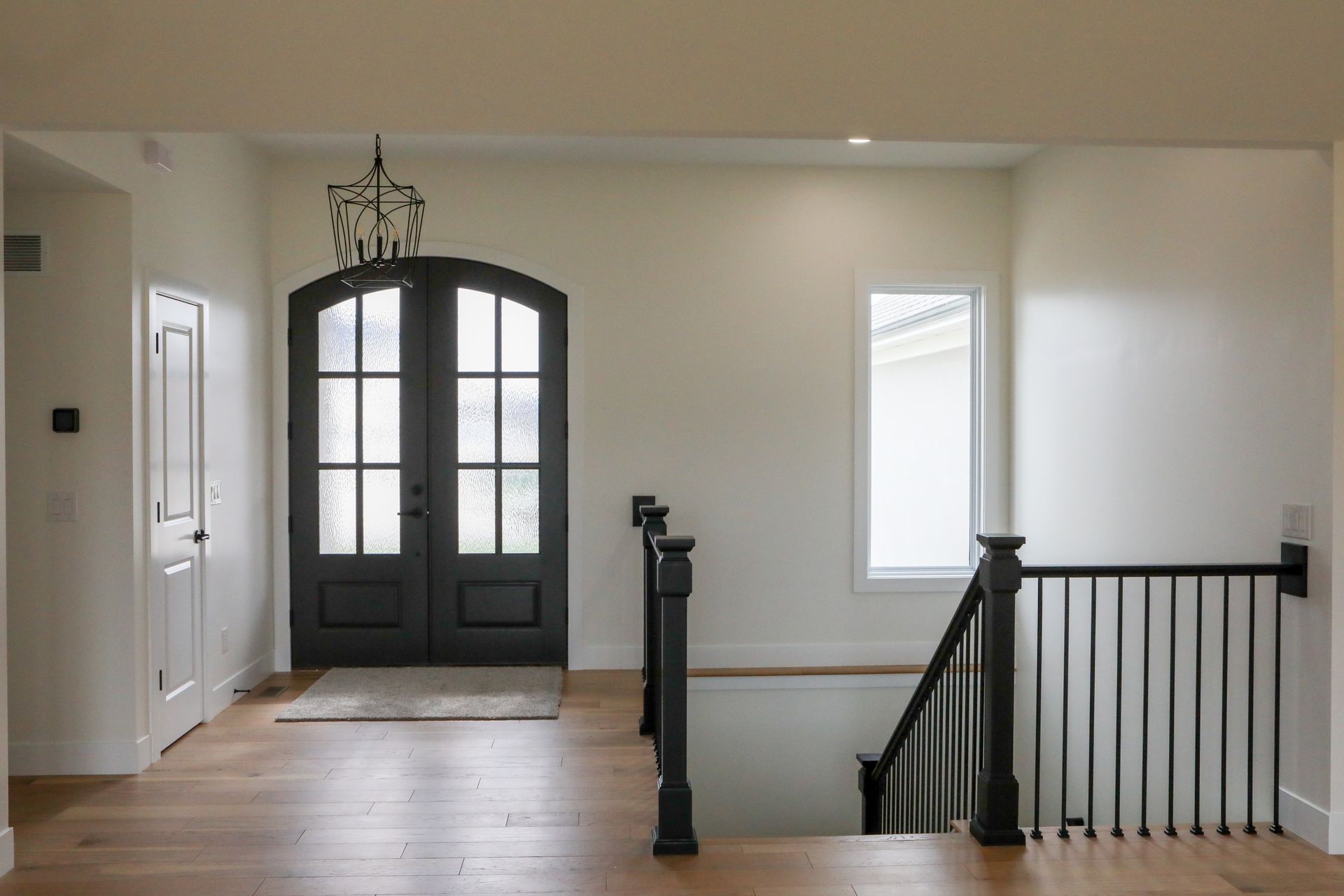 Bright foyer with black double front doors, white walls, wood floors, and a staircase railing.