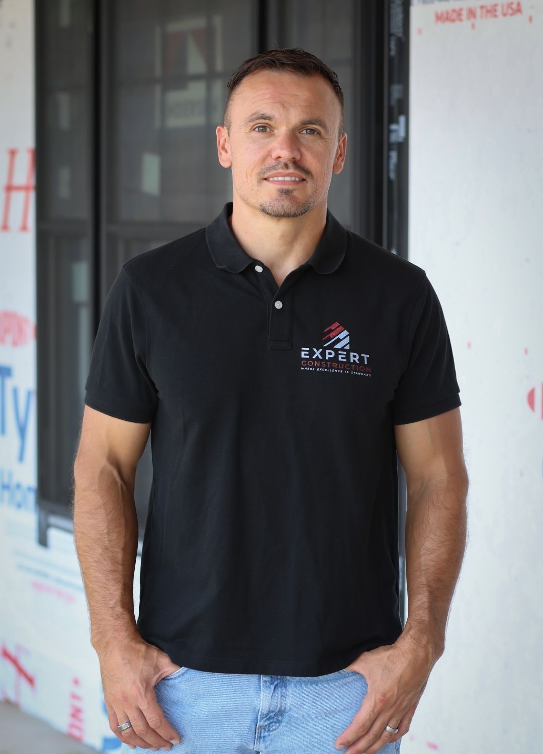 Man in a black polo shirt and jeans standing indoors by a wall.