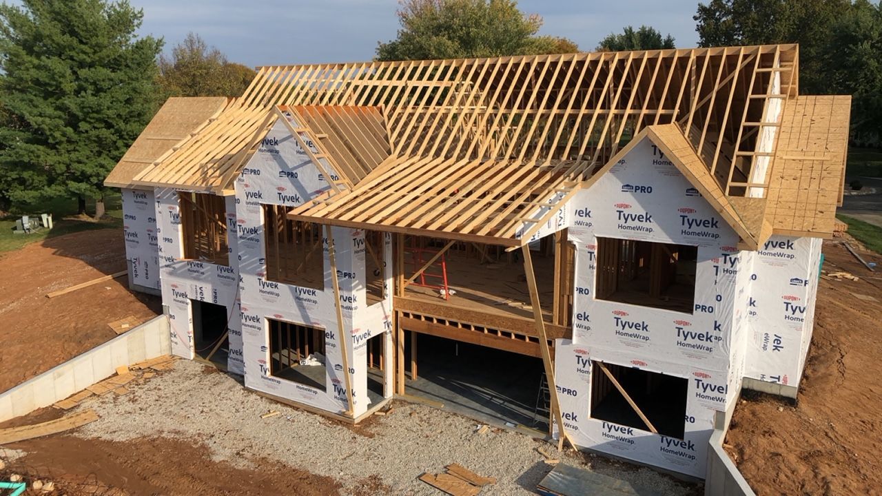 Partially built two-story house with exposed roof framing and white house wrap on a sunny day