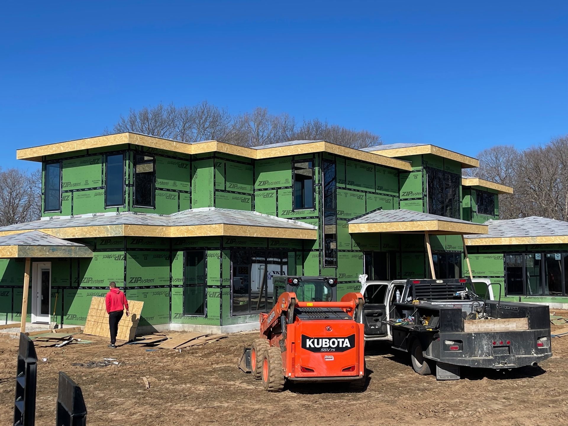 Green two-story house under construction with workers and a Kubota loader in front.