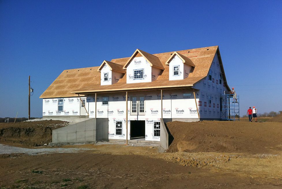 House under construction with white siding and a tan roof on a bare dirt lot, under a clear blue sky