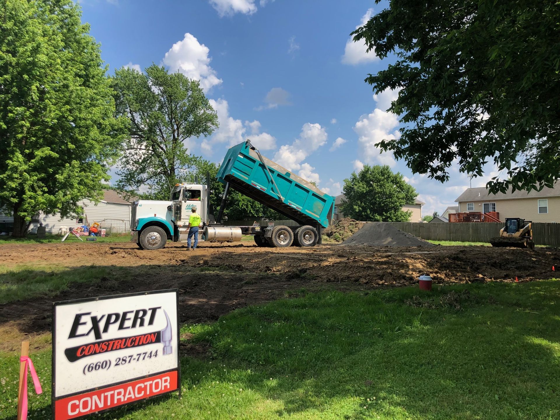Dump truck unloading dirt at a construction site with an Expert Contractors sign in the foreground