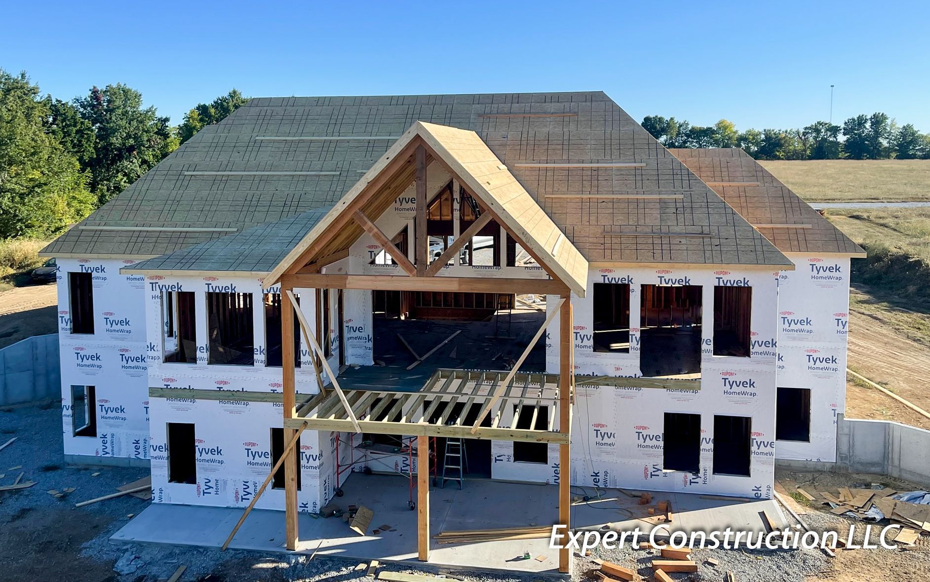 House under construction with unfinished roof, exposed framing, and white exterior wrapping at a wooded site