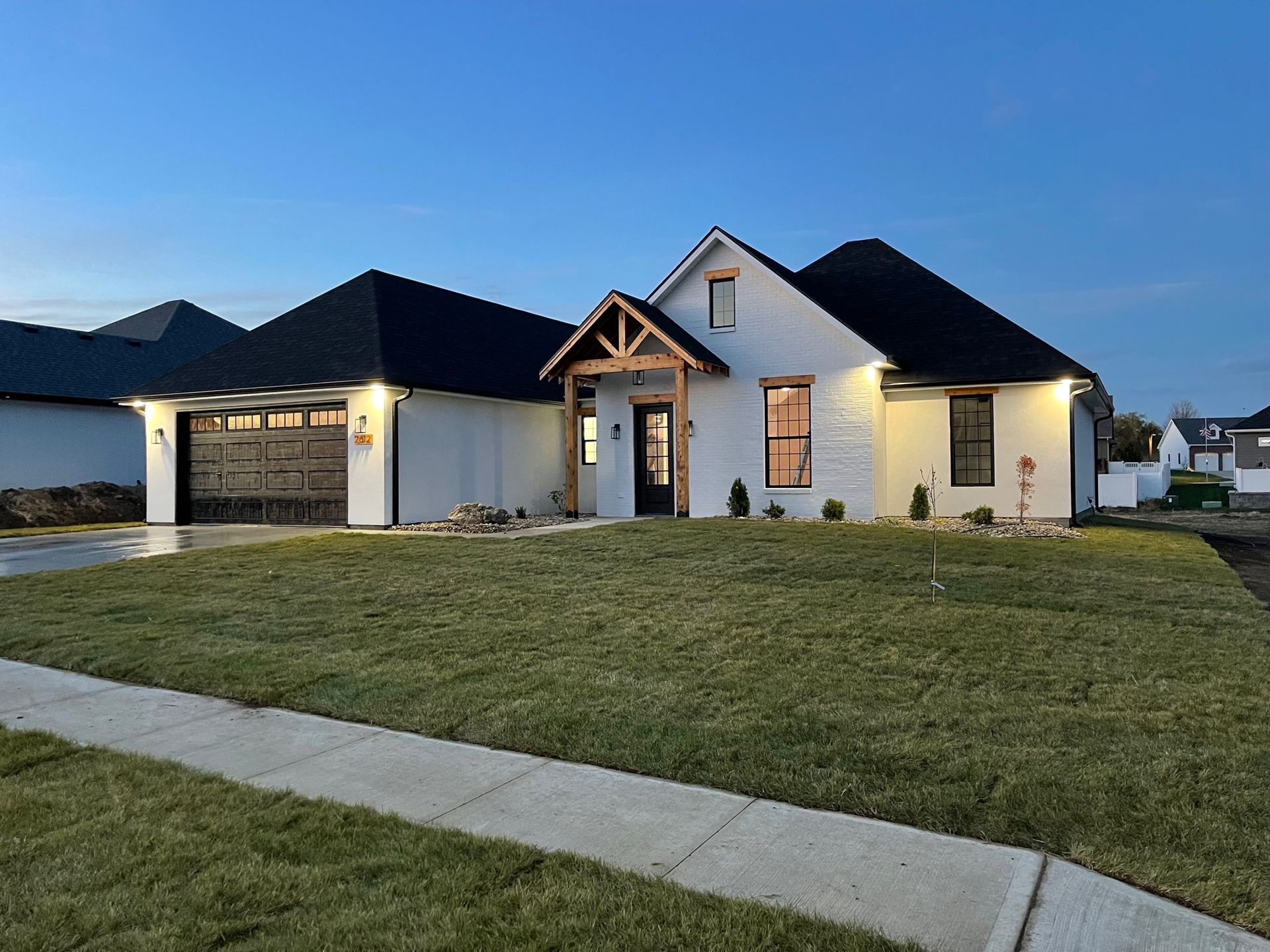Suburban house with dark roof, lit front porch, and a large green lawn at dusk