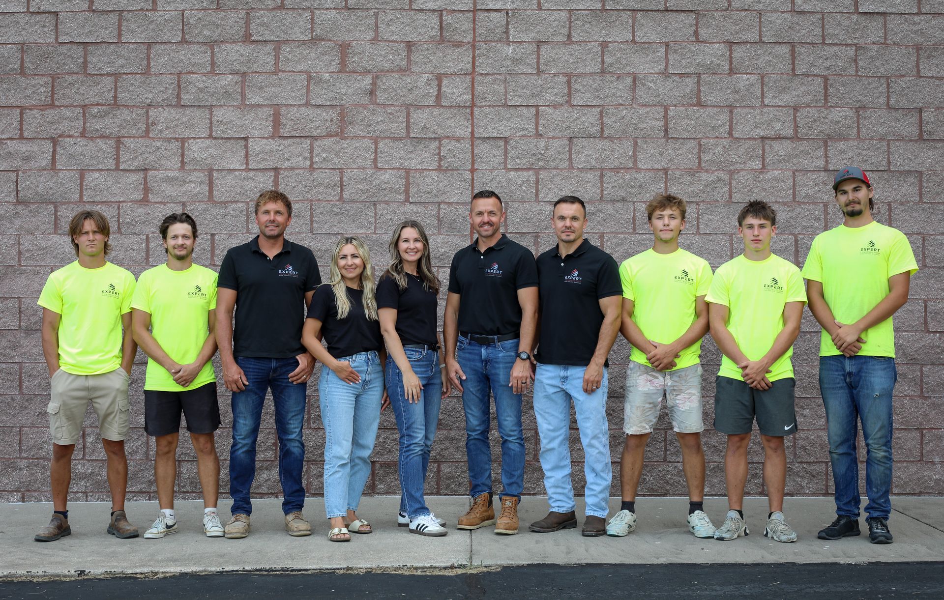 A group of people standing in a row against a stone wall, several wearing neon yellow shirts and jeans.
