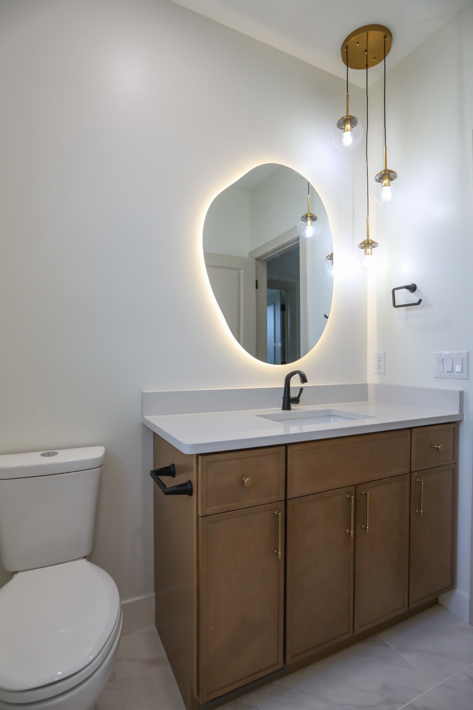 Small bathroom with a lit oval mirror, wood vanity, toilet, and pendant lights.