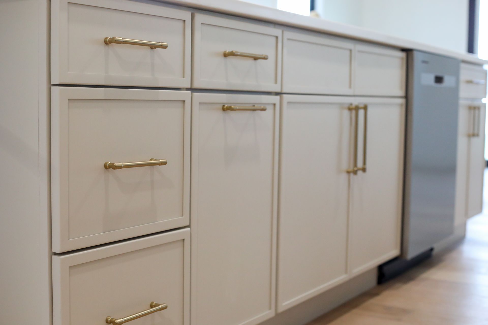 White kitchen cabinets with brass handles and a stainless steel dishwasher along a light wood floor.