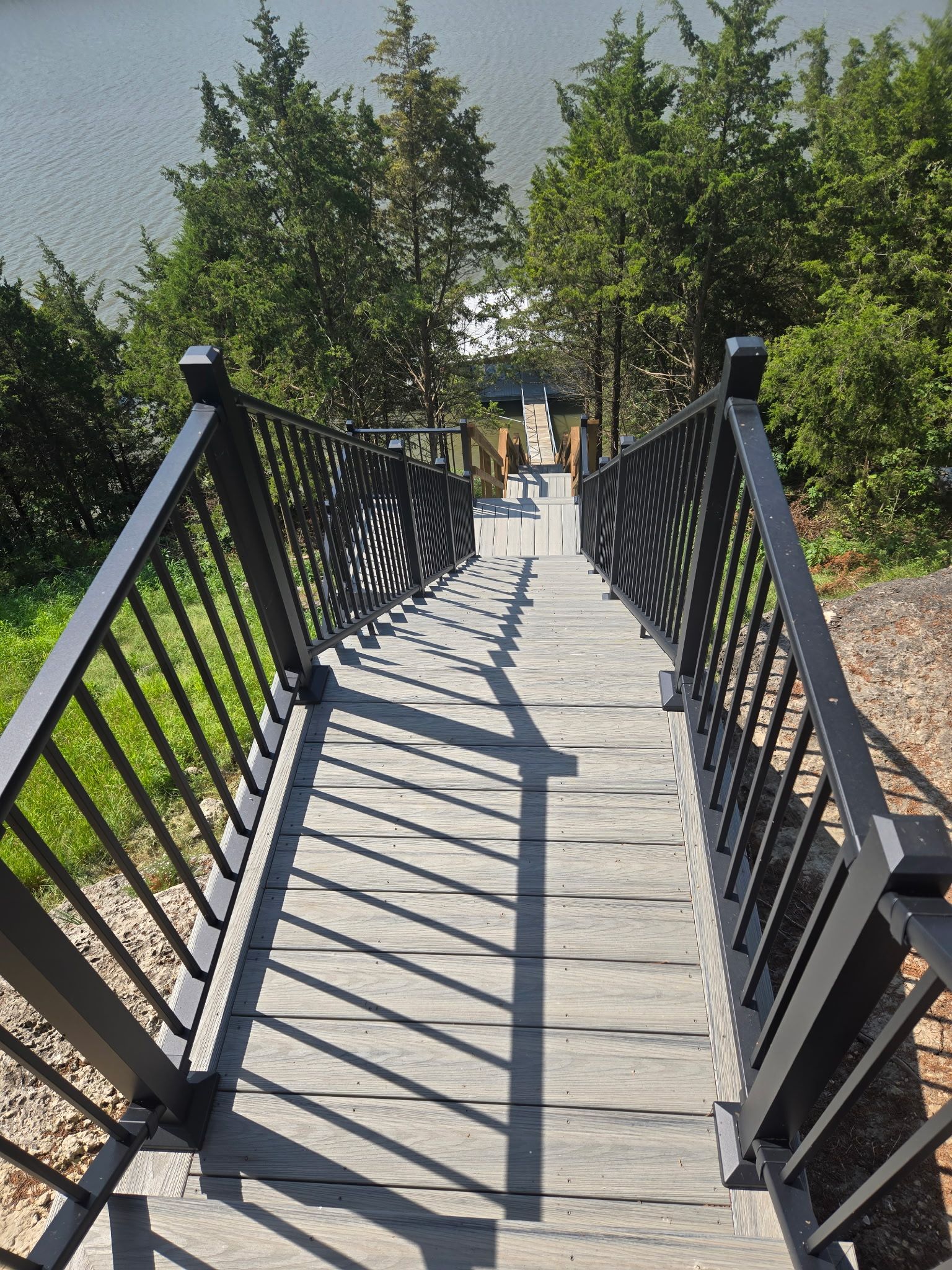 Narrow suspension bridge with black railings over water, leading toward trees in a sunny park
