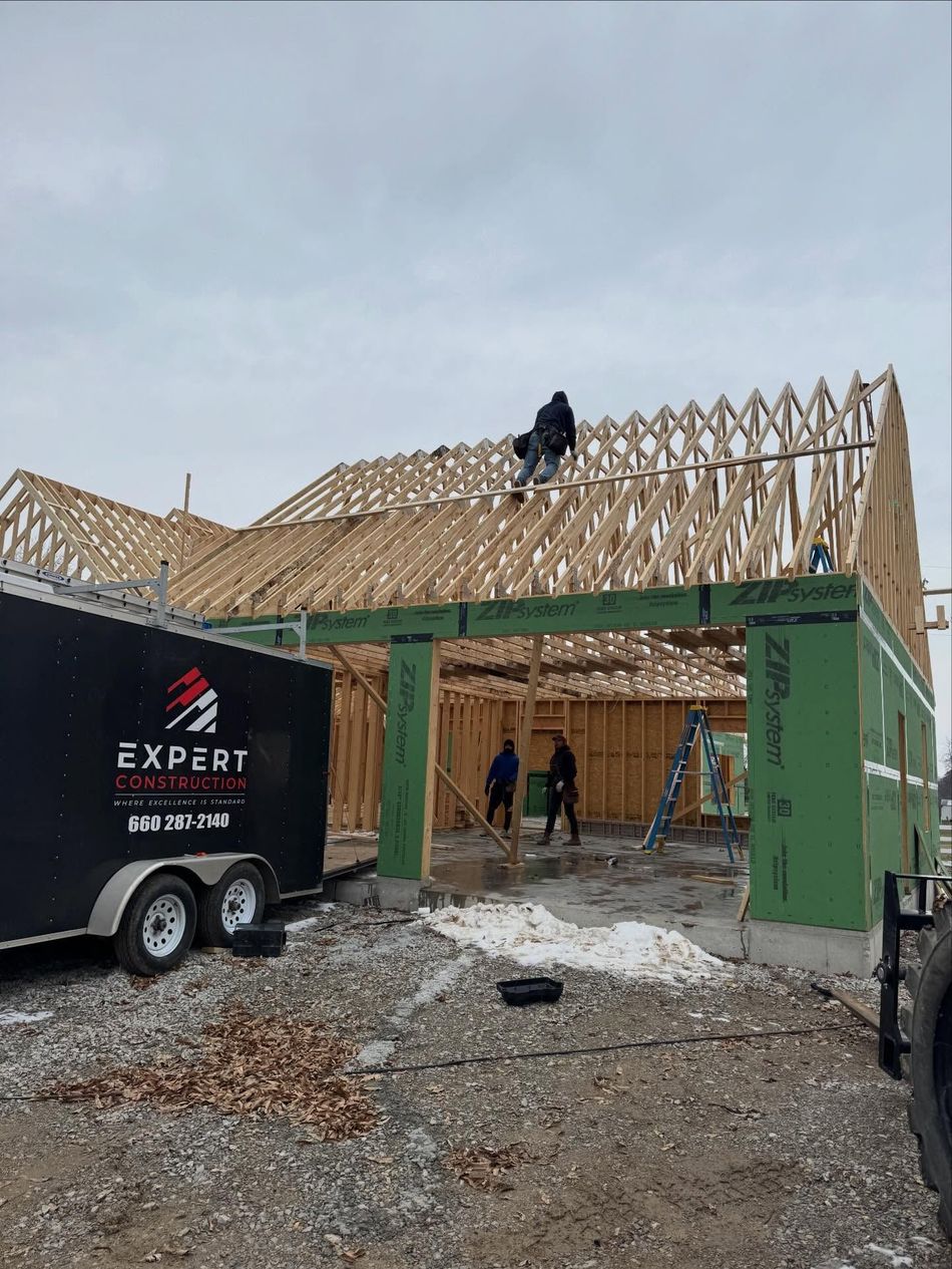 House under construction with exposed wooden roof framing, workers on site, and a black trailer in front.