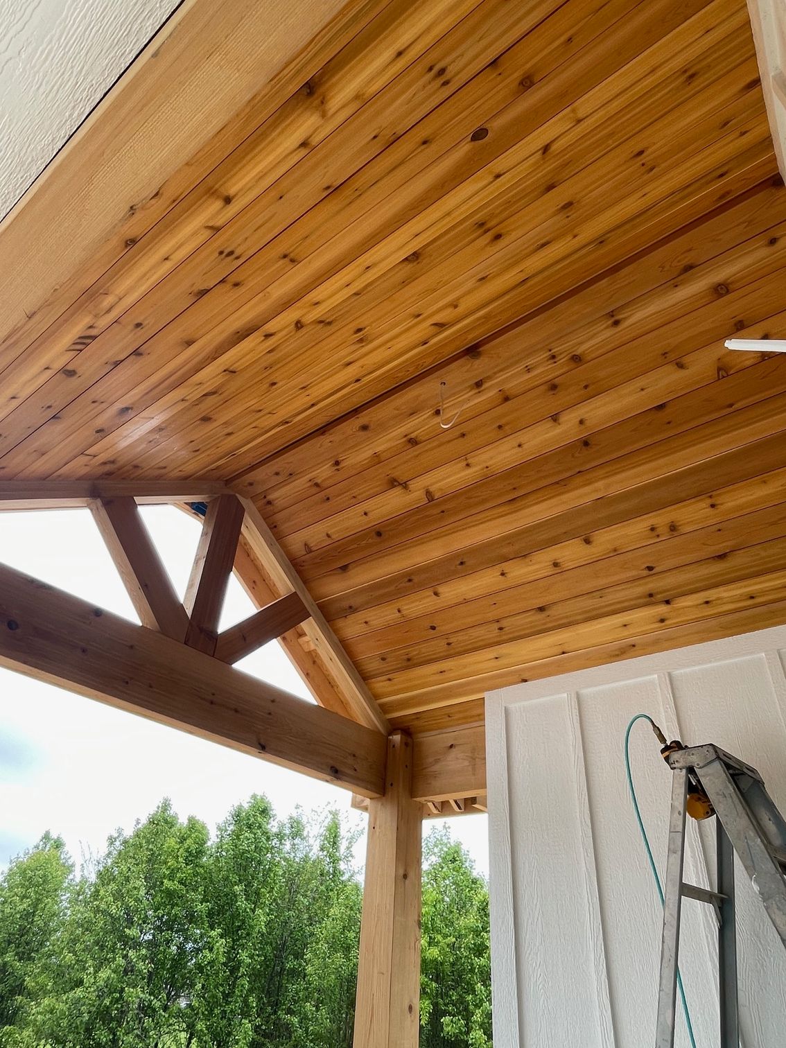 Wooden porch ceiling under construction, with beams, ladder, and trees visible outside.