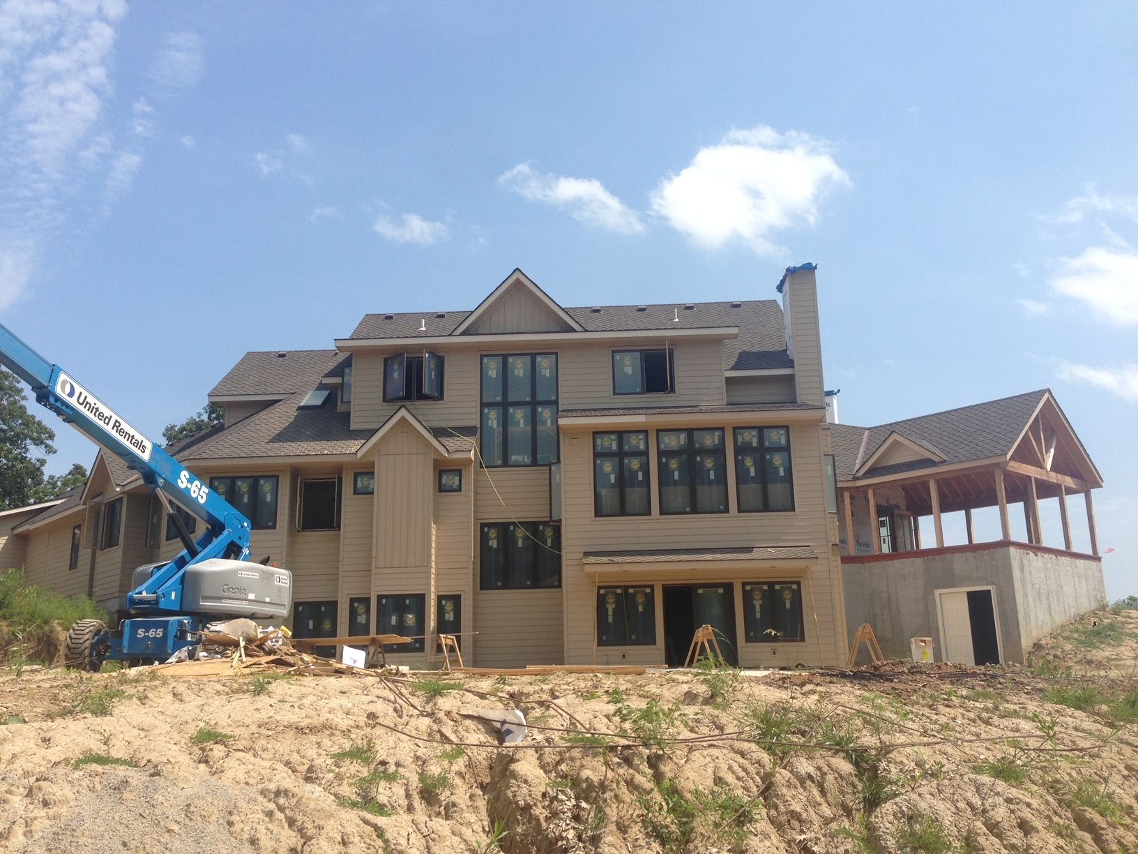 Partially built two-story house on a dirt lot with a blue construction lift under a bright sky