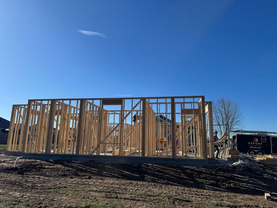 Wood-framed house under construction on a dirt lot beneath a clear blue sky.