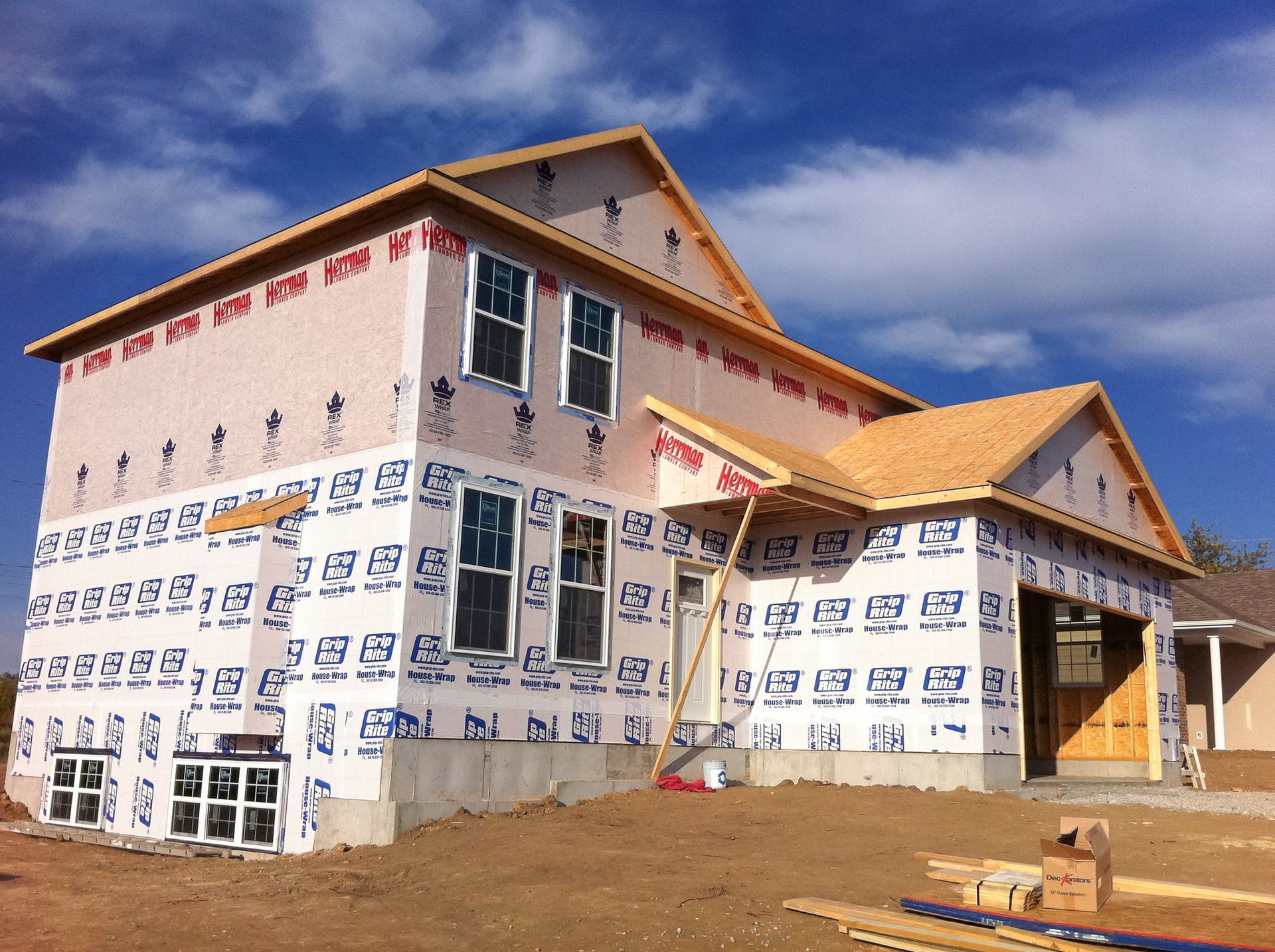Two-story house under construction with wrapped exterior and a dirt lot under a blue sky