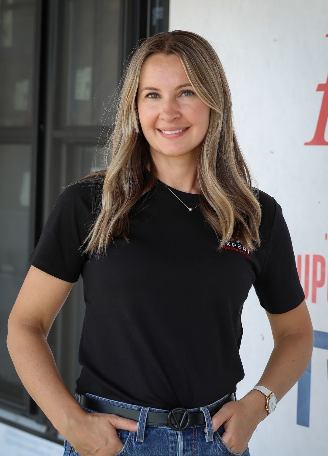 Smiling woman in a black shirt and jeans standing indoors with hands on hips, beside a wall and windows.