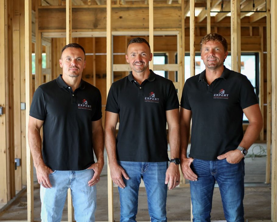 Three men in black polo shirts standing inside an unfinished wooden building frame.
