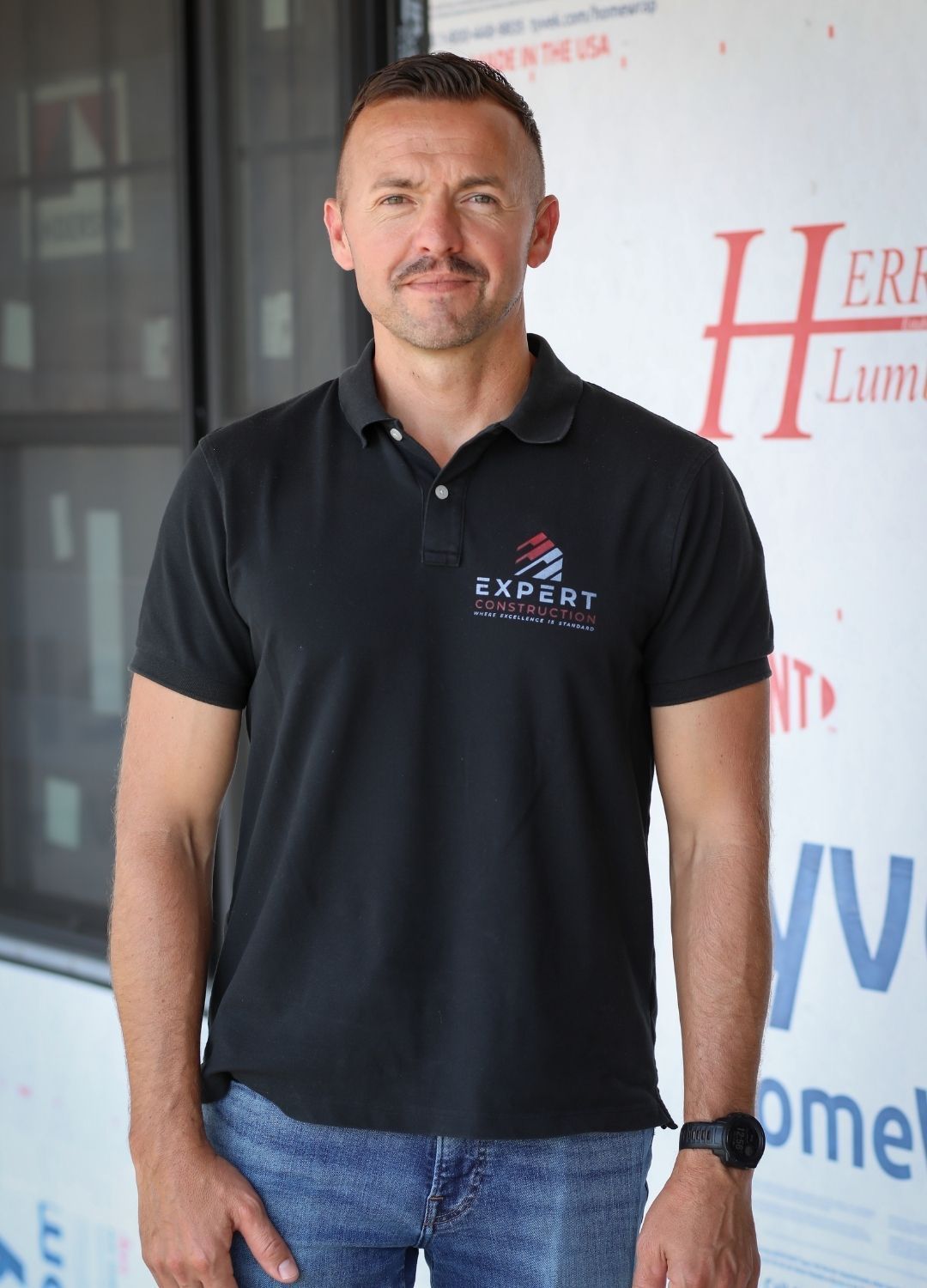 Man in black polo shirt and jeans standing indoors beside a branded wall banner