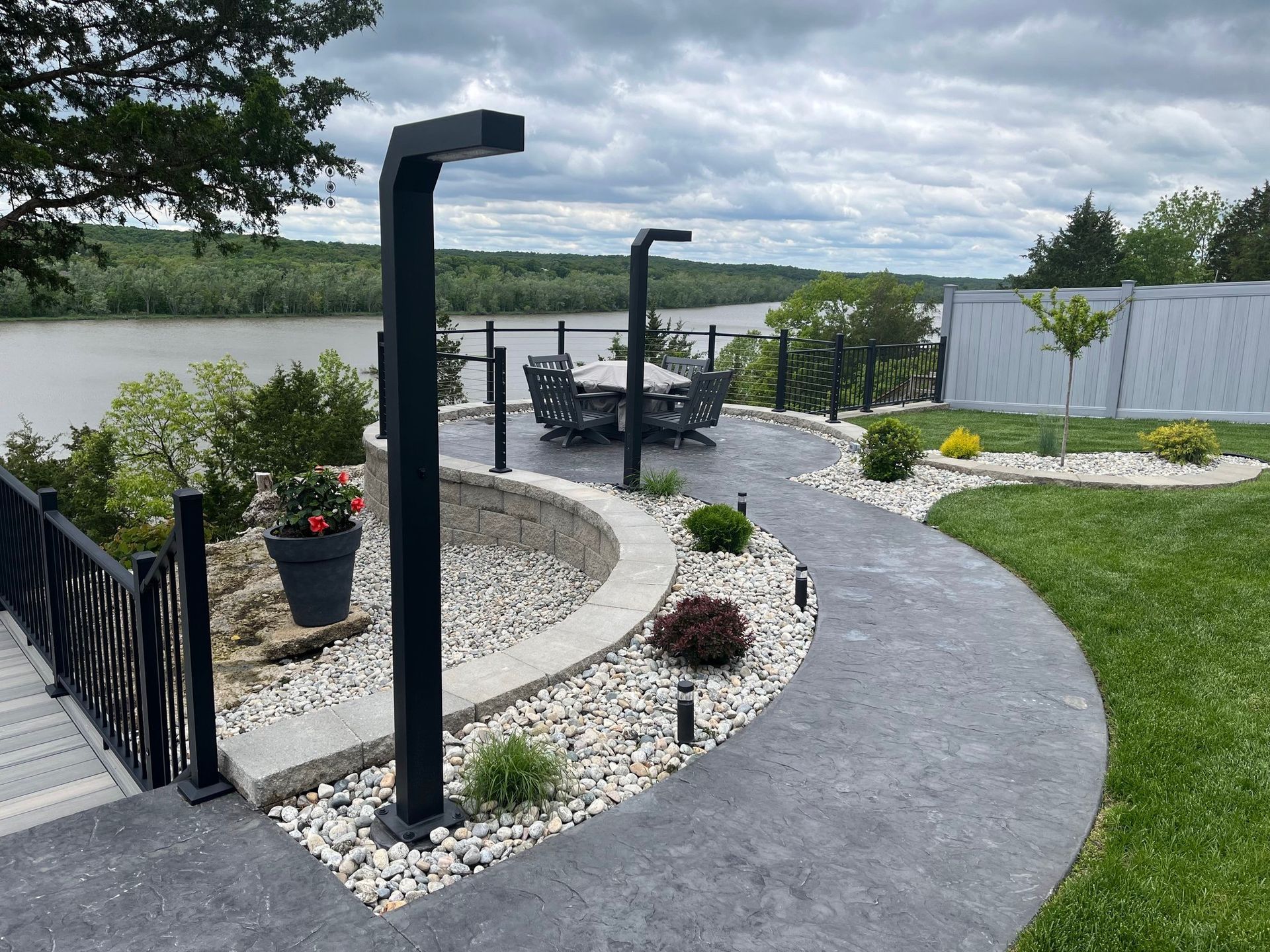 Curved stone walkway by a lakeside patio with black posts, potted flowers, and a table near a railing