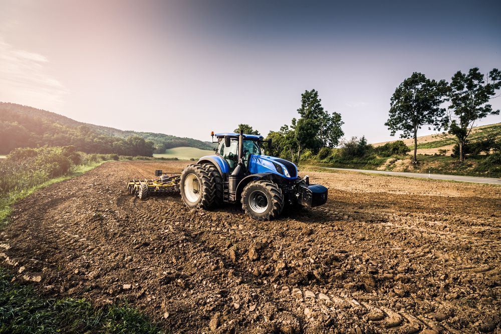 Blue tractor plowing a dry field under a bright sky with hills in the background — Bundaberg Field Service in Bundaberg East, QLD