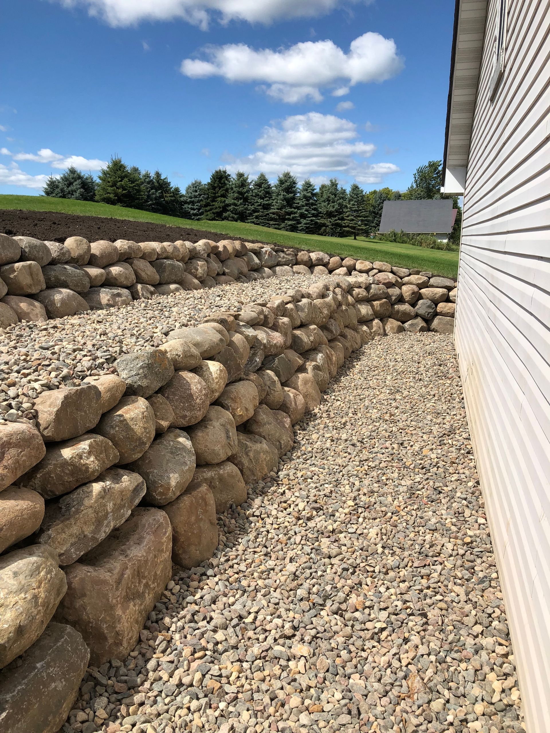 Terraced stone retaining walls beside a gravel path, with a house wall and blue sky in the background