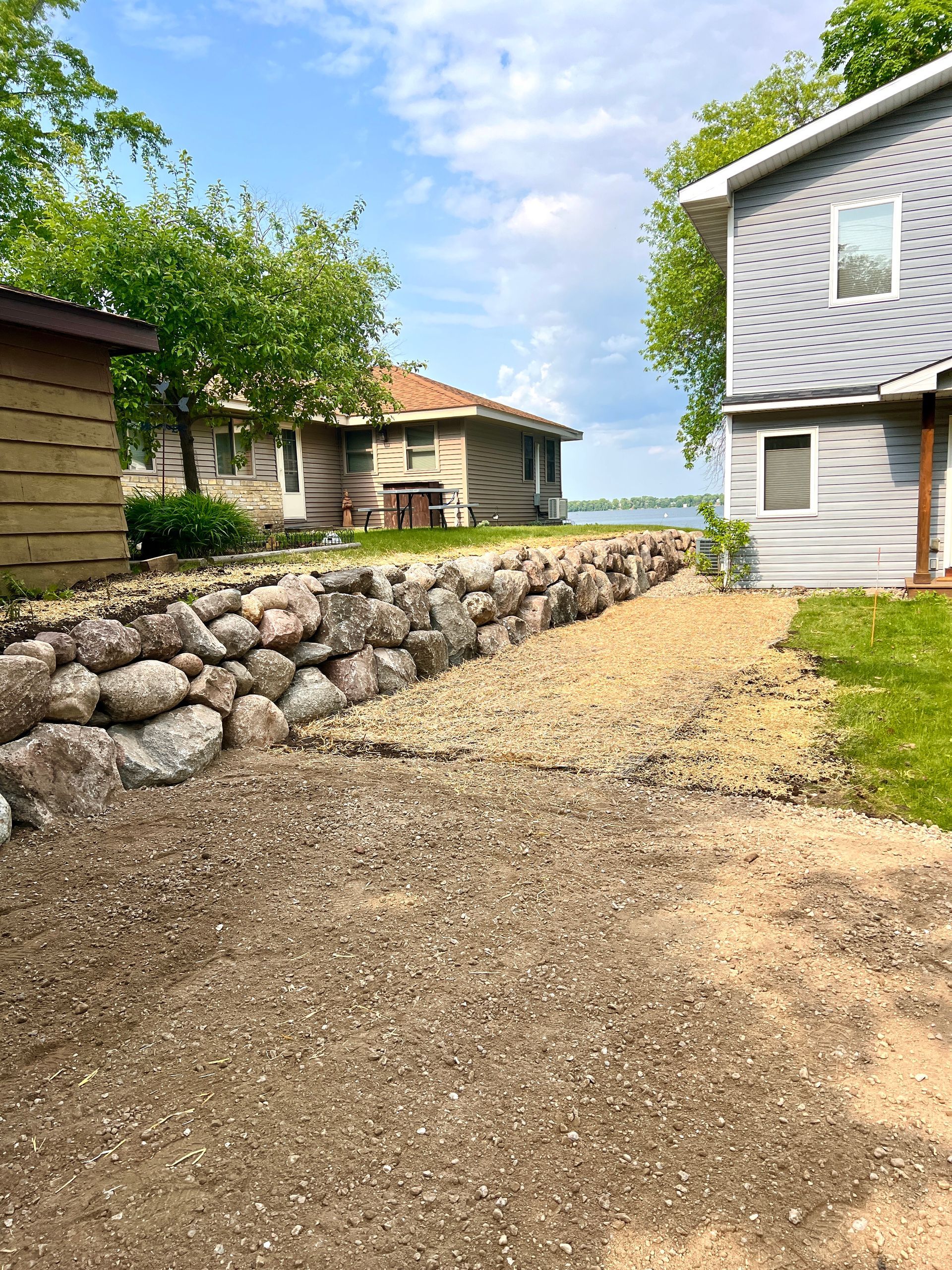Gravel path between houses leading to a lake, with stone retaining wall and green trees under a blue sky.