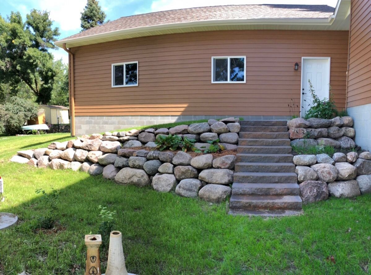 House with stone retaining wall and central steps leading to a white door, set on a grassy lawn.