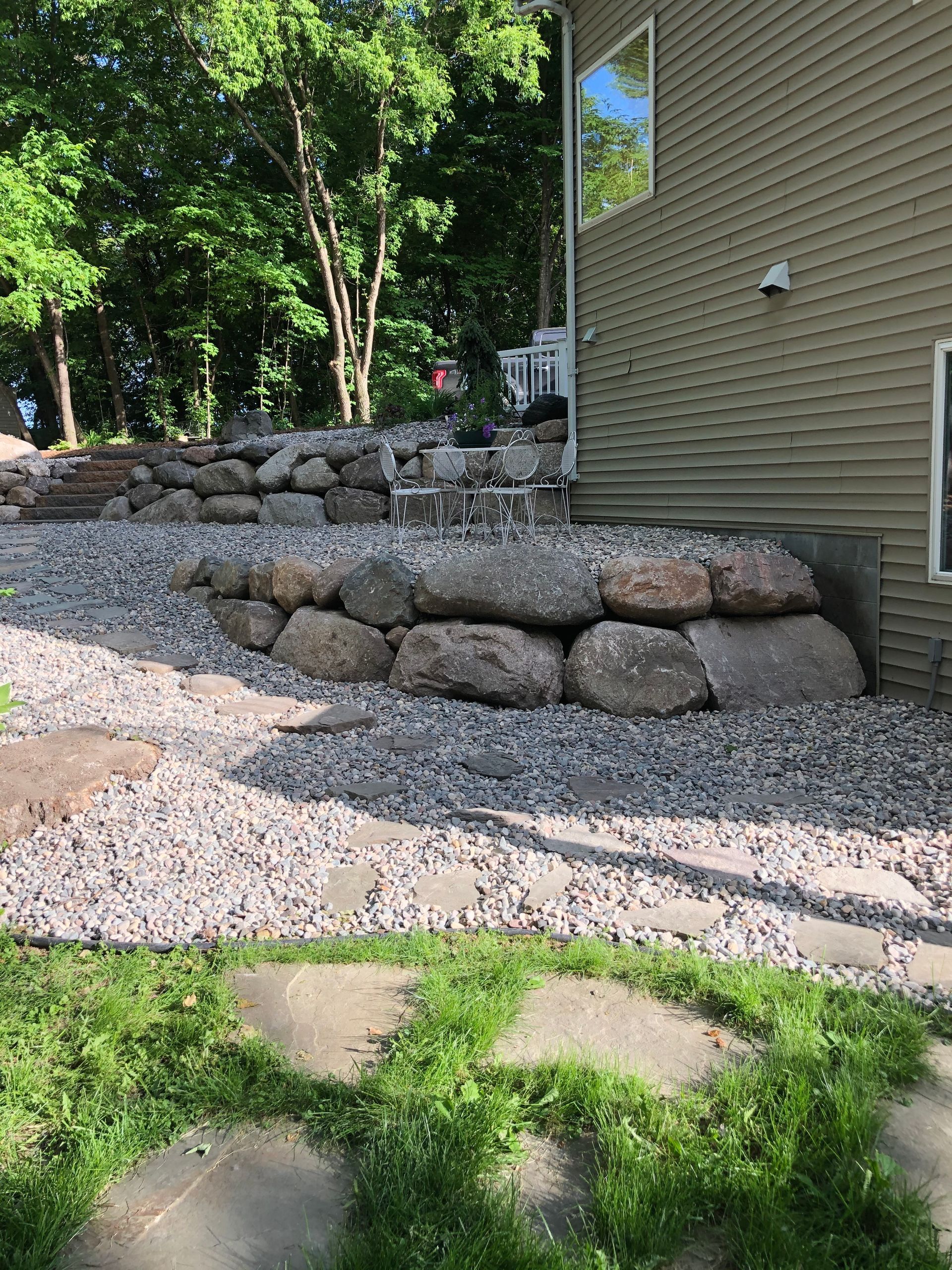 Stone retaining wall beside beige house with gravel path and green lawn in front
