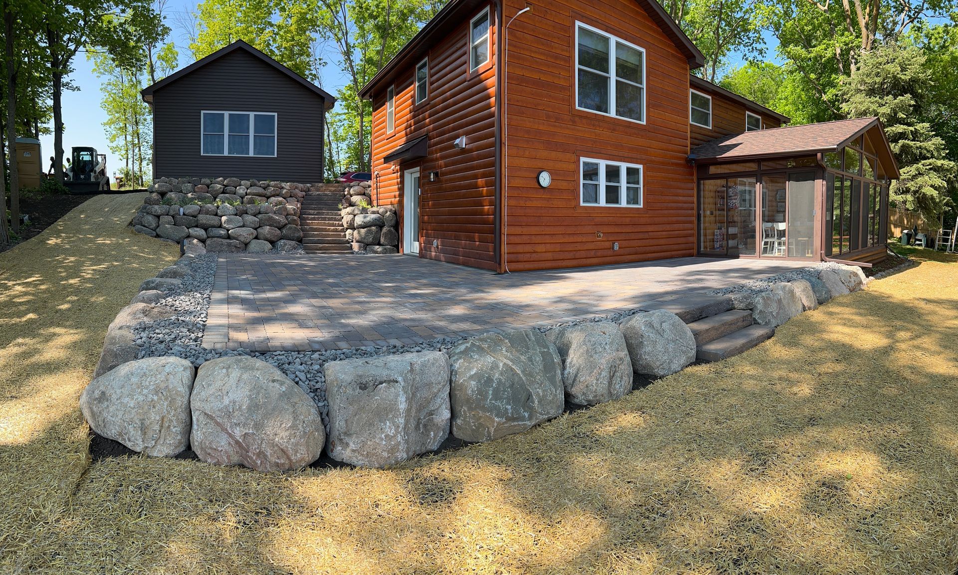 Wooded yard with a red wooden cabin, gray shed, and stone retaining wall on a sloped lawn.