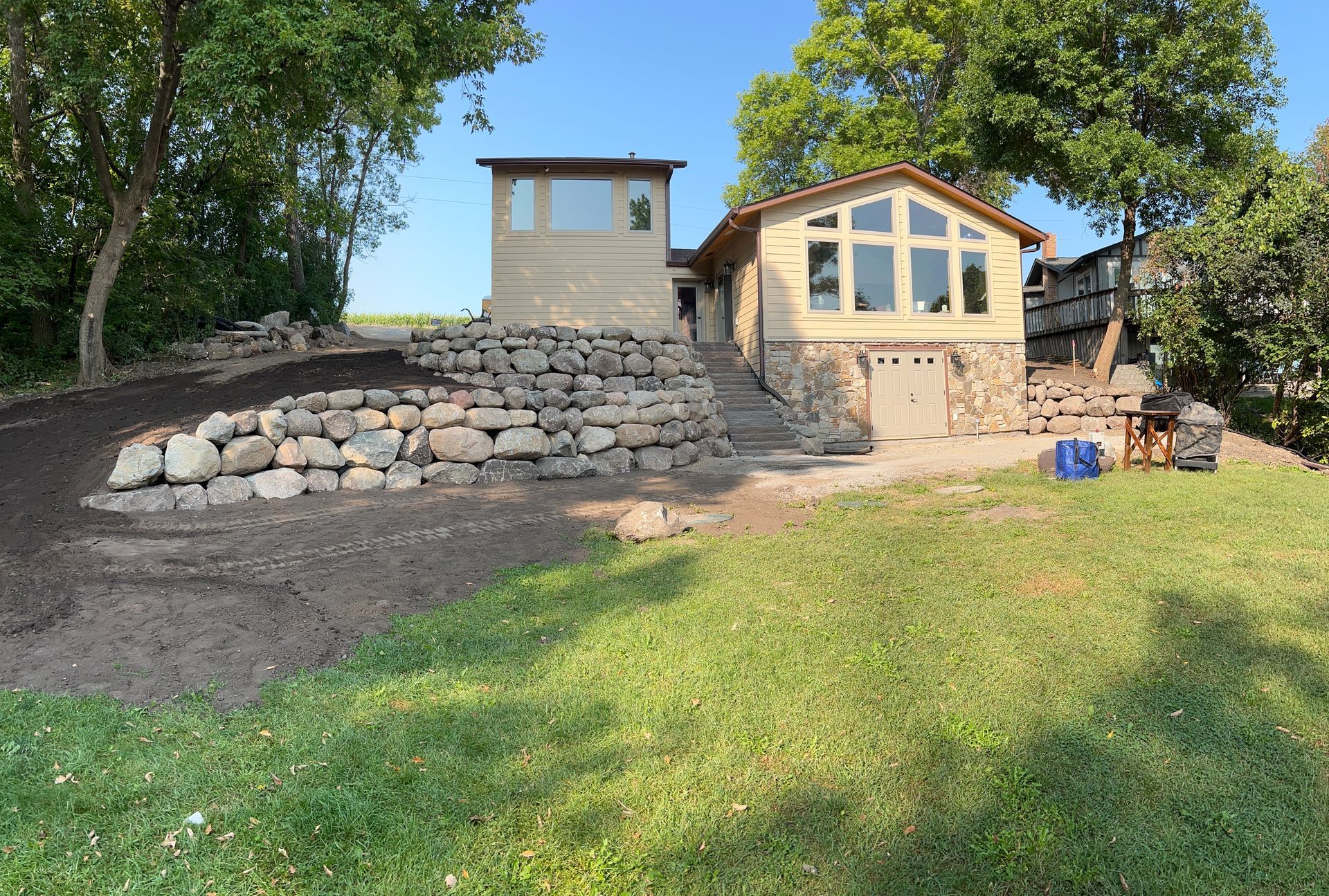 Stone retaining wall and steps leading to a small house with large windows, beside a grassy yard and trees.