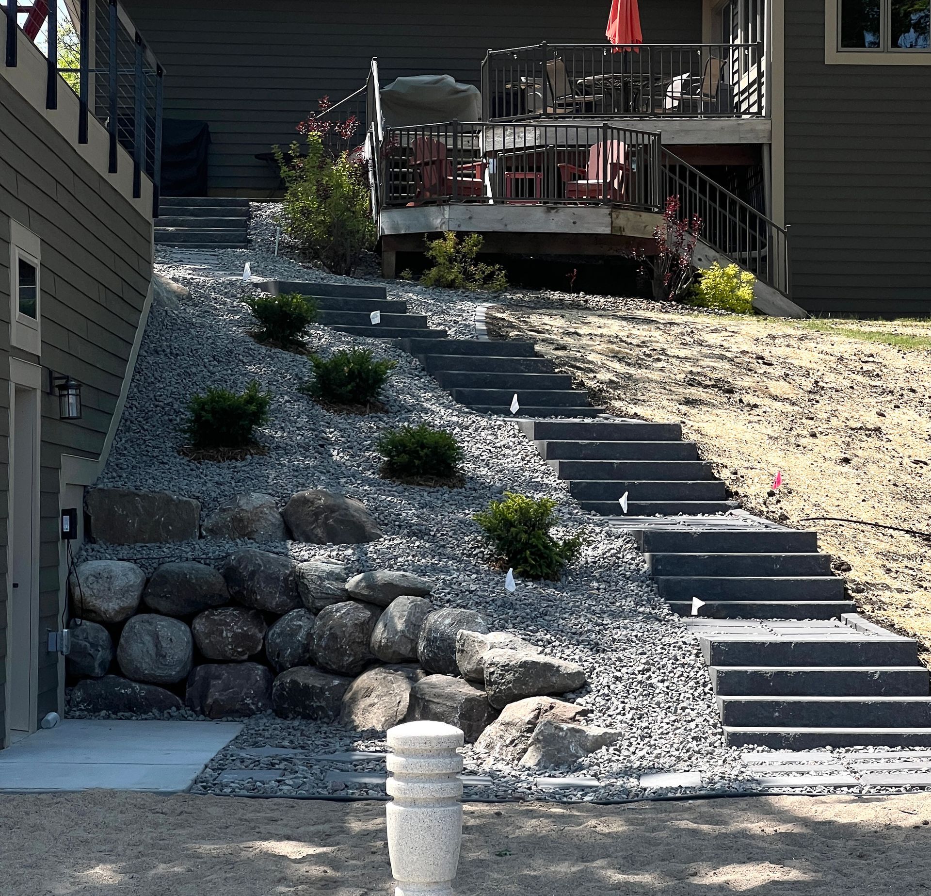 Steep landscaped outdoor staircase with stone retaining wall and deck patio above on a residential hillside