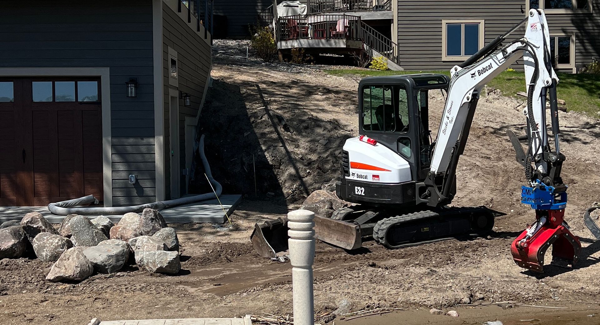 Compact excavator digging dirt beside a house, with rocks and a red attachment in a construction yard.