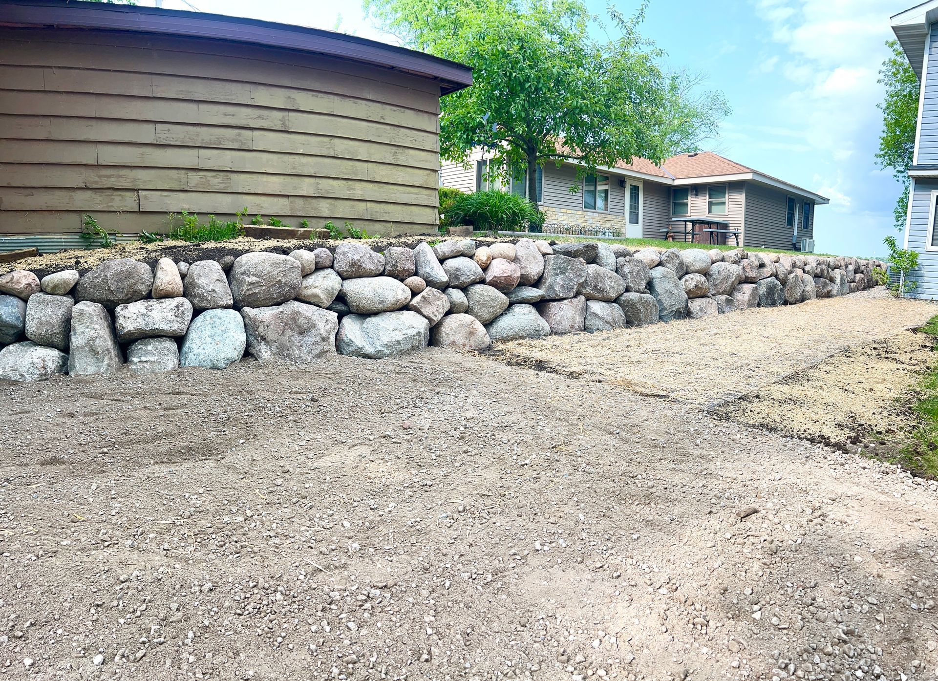 Rock retaining wall beside a gravel driveway with houses and trees in the background