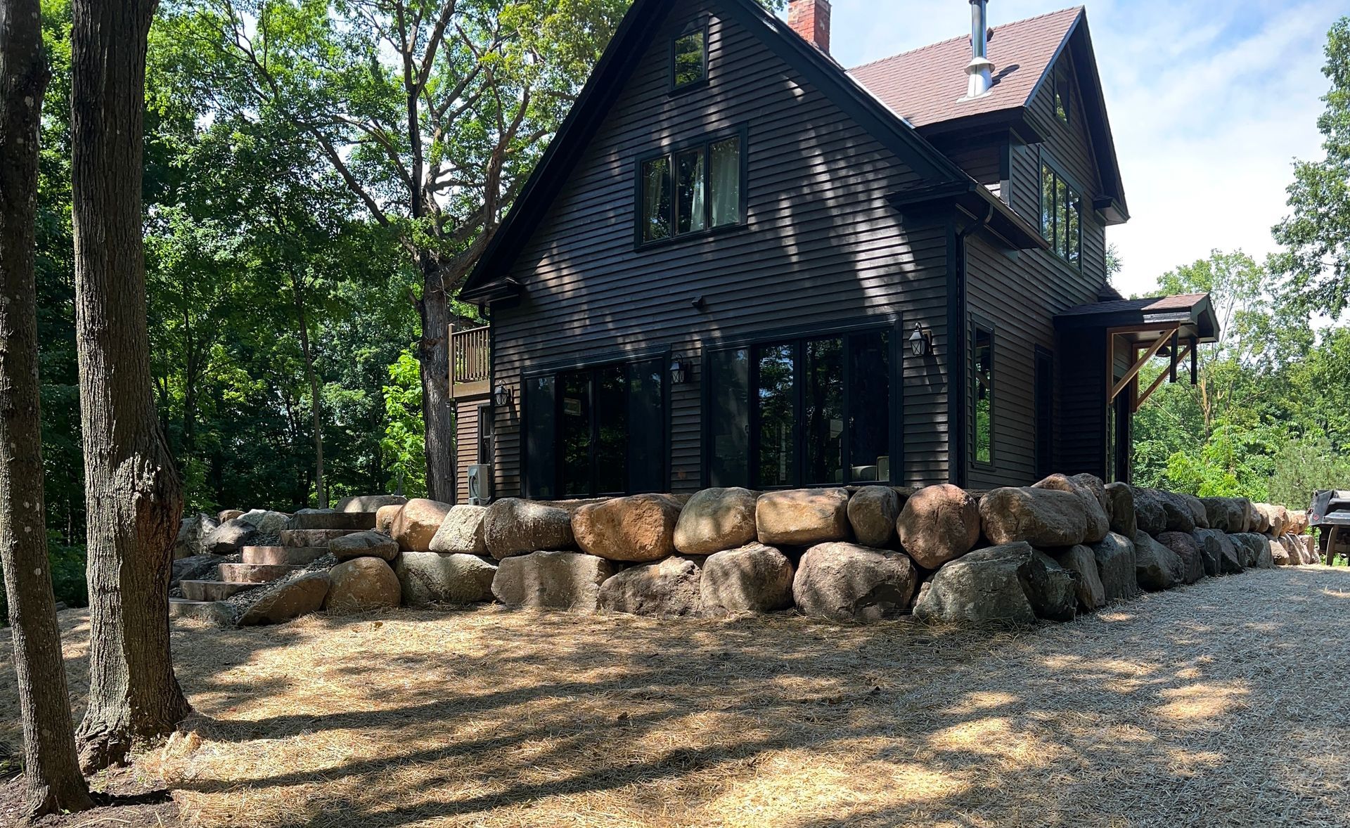 Dark wooden house with stone foundation surrounded by trees and a gravel yard