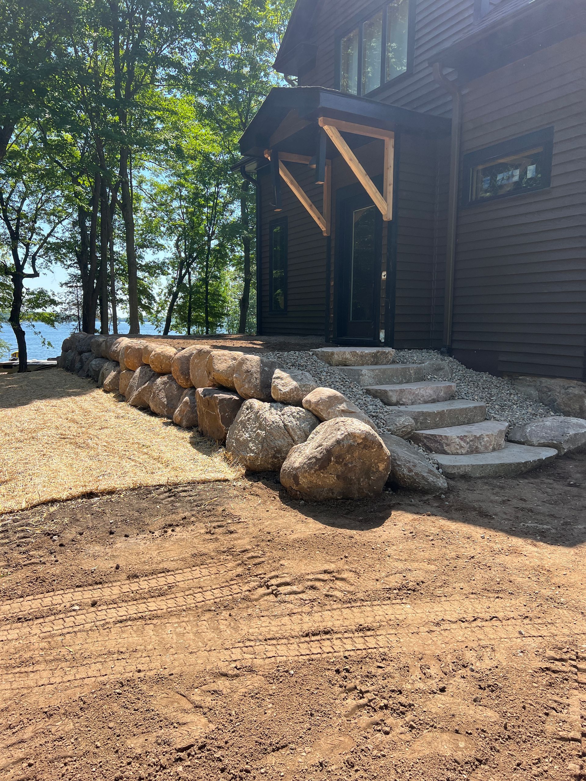Stone steps beside a dark wooden cabin, with a row of large boulders and trees by the water