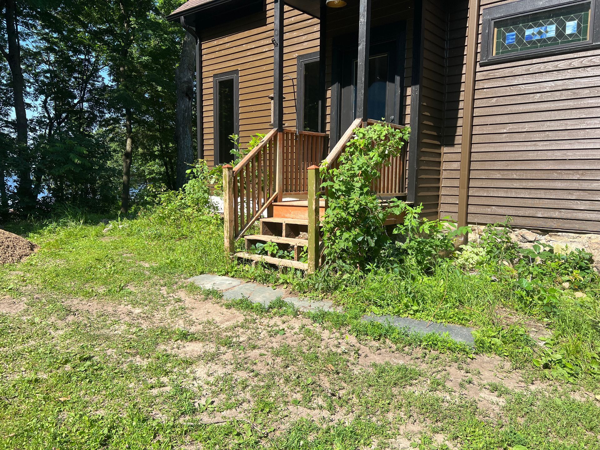 Small wooden porch with stairs on a brown house, surrounded by greenery and a grassy yard.