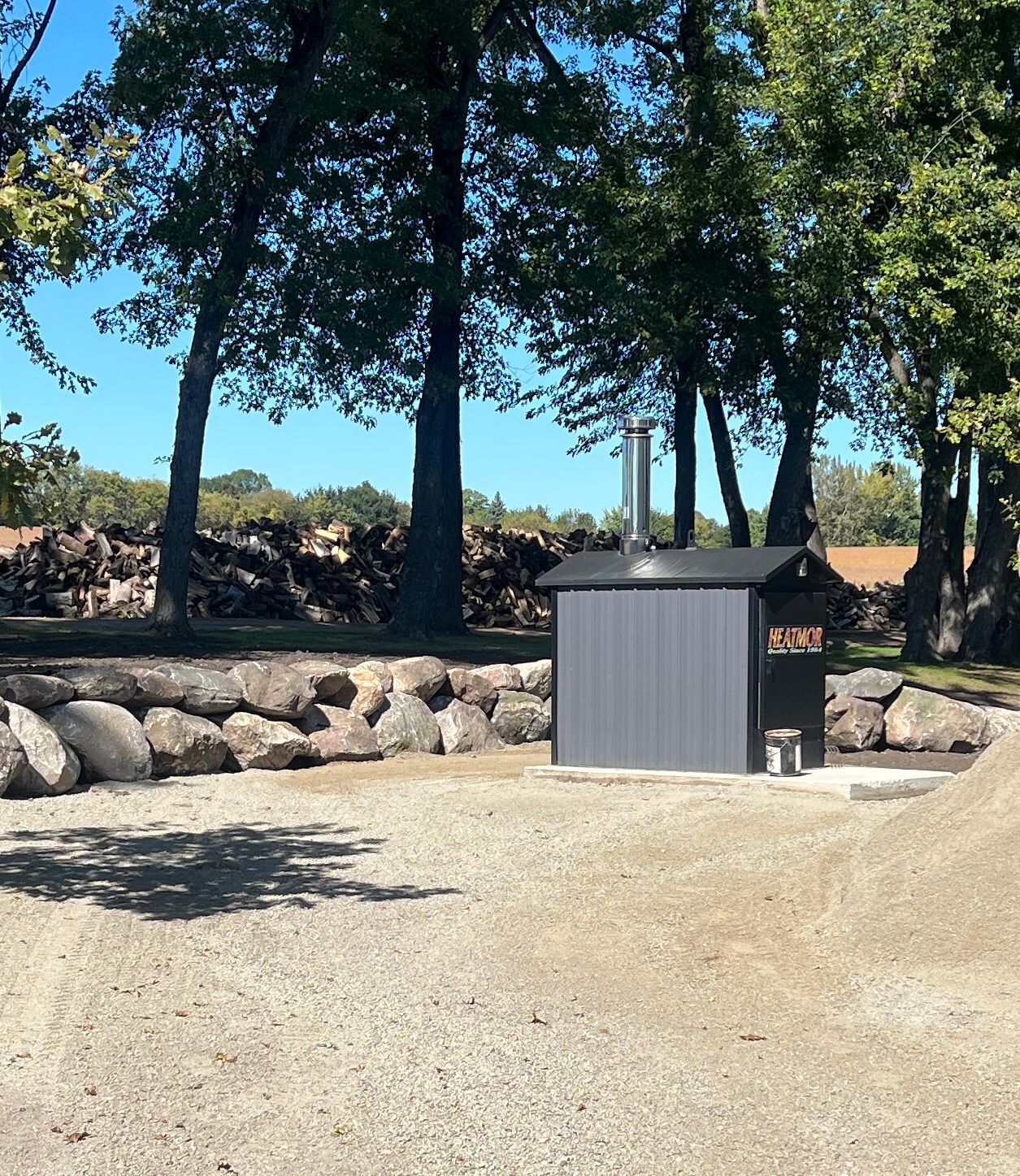 Outdoor gravel area with trees, rocks, and a gray utility box near a small sign.
