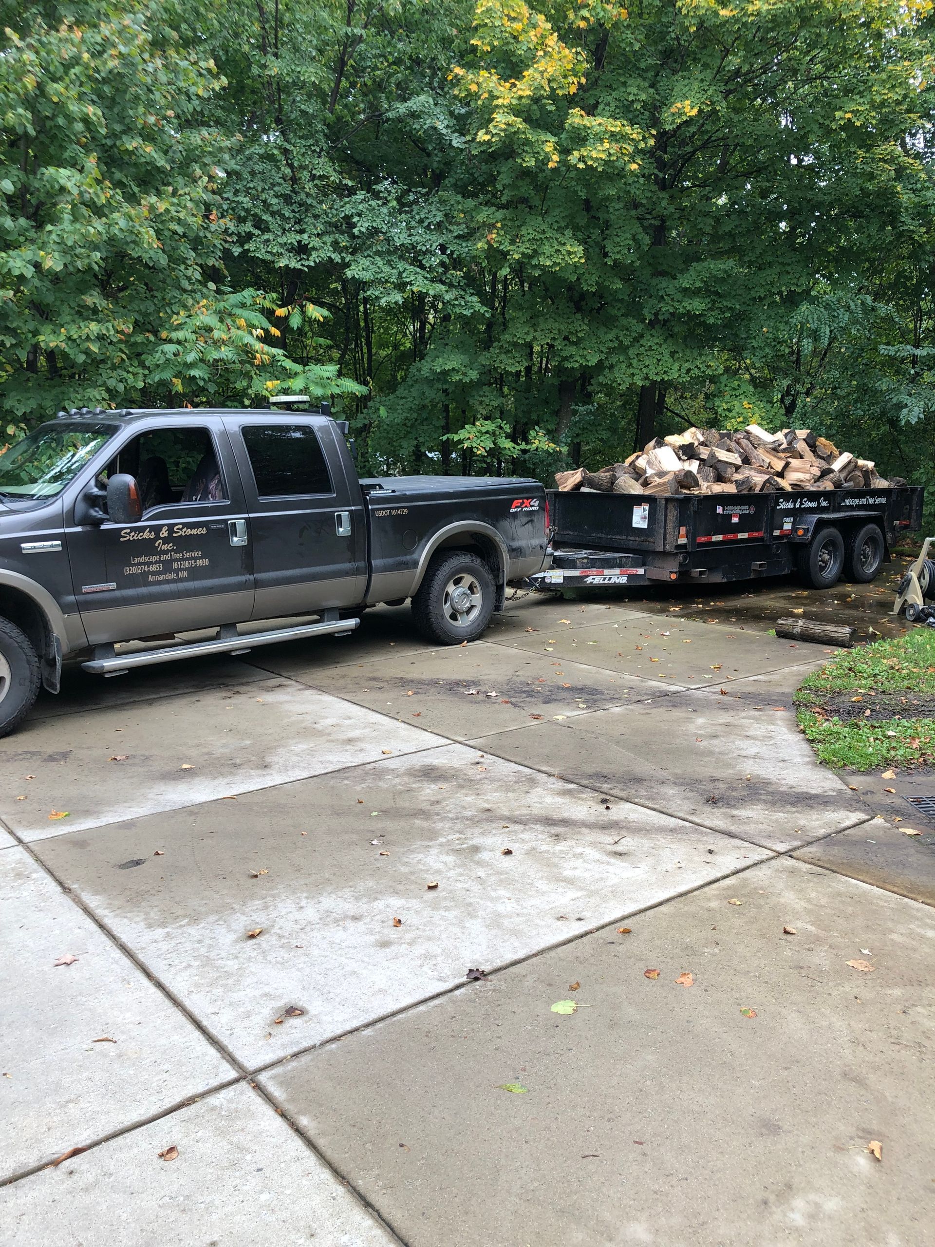 Black pickup truck towing a flatbed trailer piled with cut branches beside a wet driveway