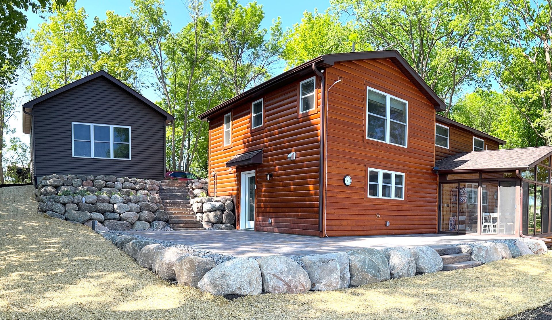 Two modern houses with dark siding and wood exterior on a rocky hillside under green trees