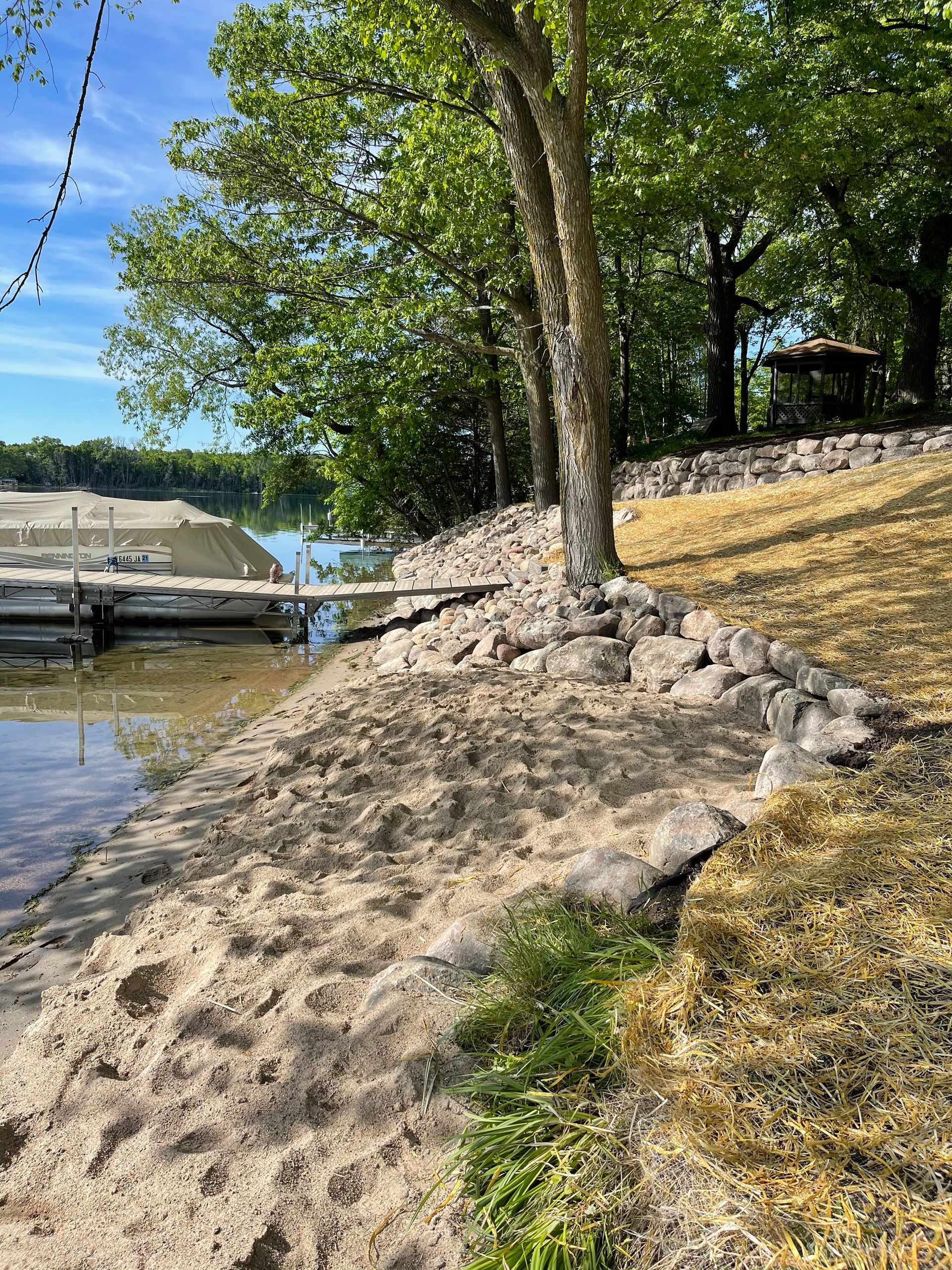 Sandy lakeshore with a tree on a rocky bank, docked boat, and calm water under a clear blue sky
