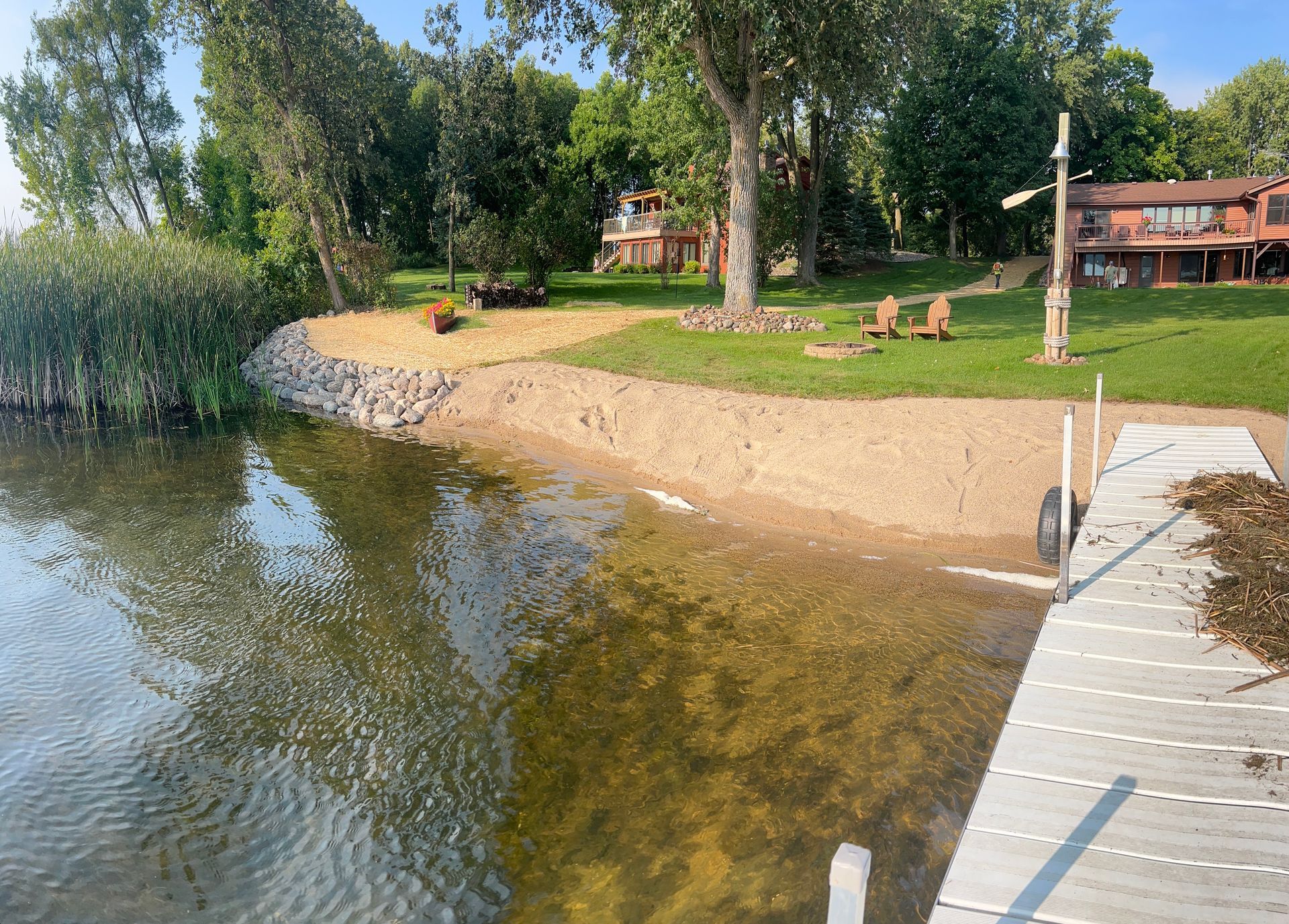 Lakeside beach with dock, trees, and a small cabin in the background on a sunny day