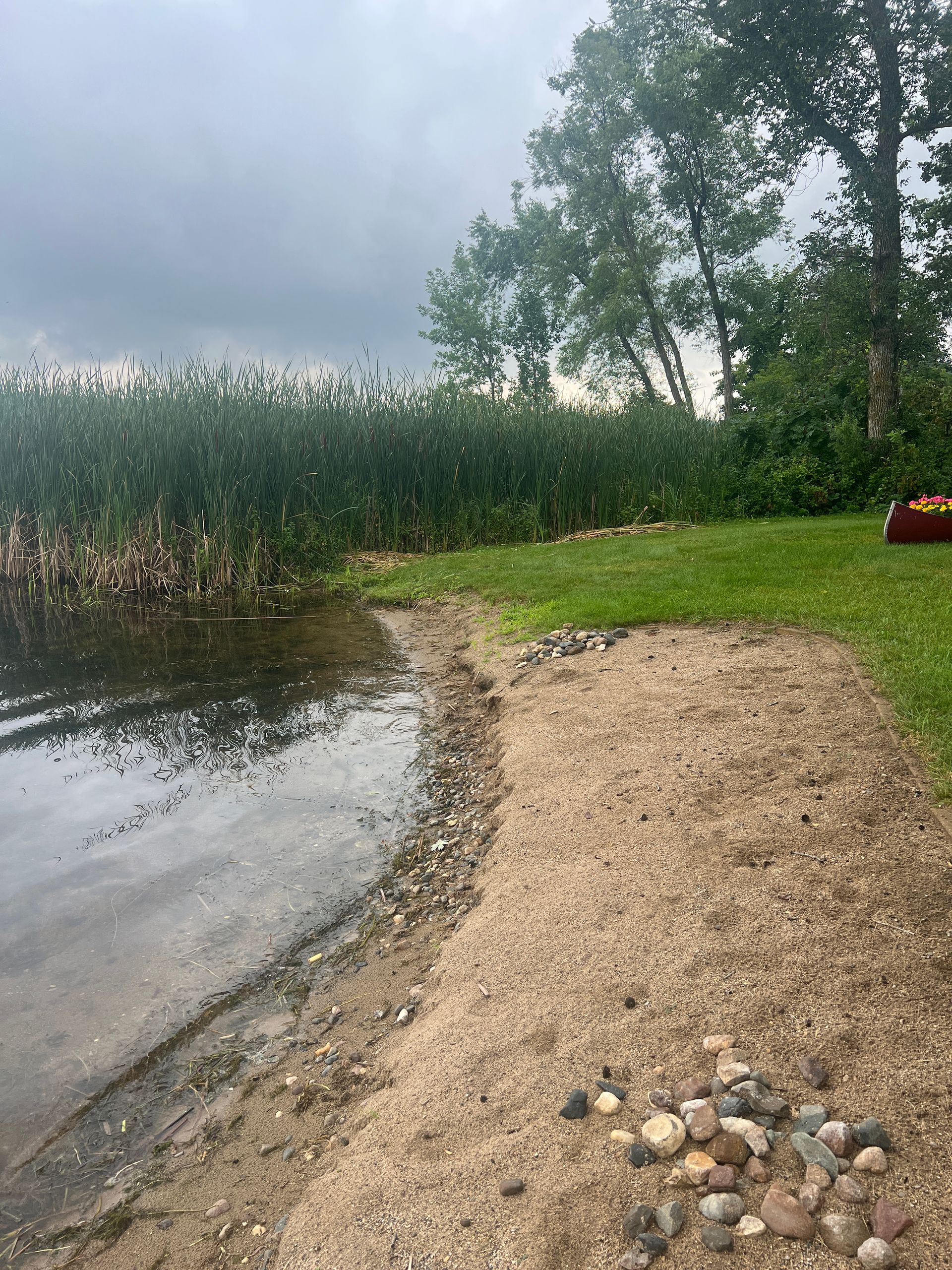 Rocky lakeshore with a gravel path, calm water, reeds, and a cloudy sky.