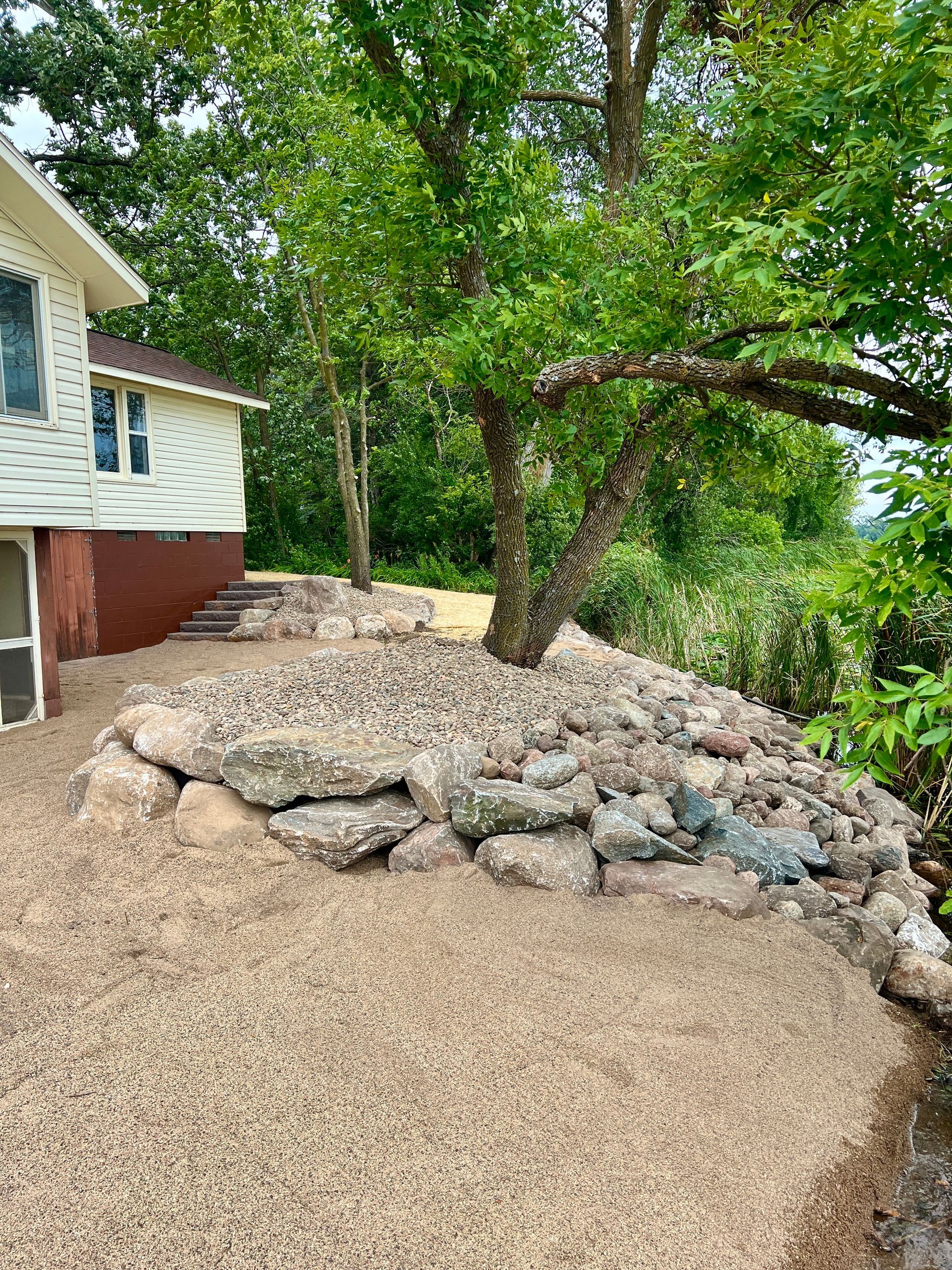 Gravel driveway beside a house with a stone retaining wall and a large tree