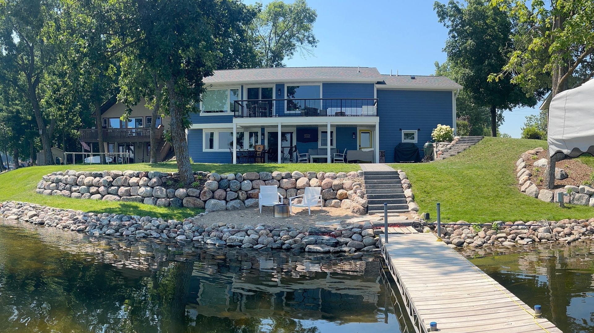 Lakeside modern house with stone retaining wall, dock, and grassy yard under a blue sky