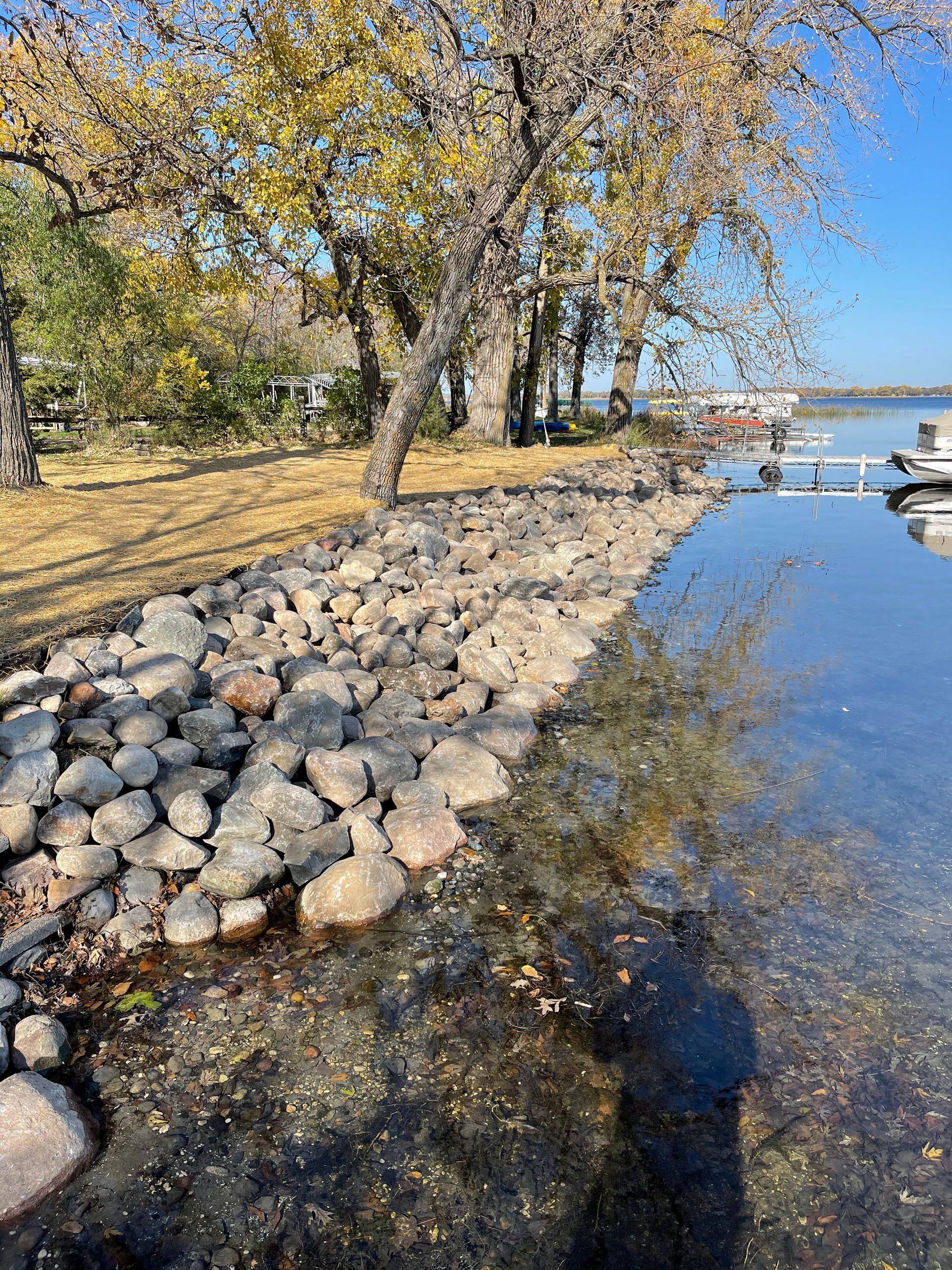 Rocky shoreline beside calm water, with autumn trees and a clear blue sky.