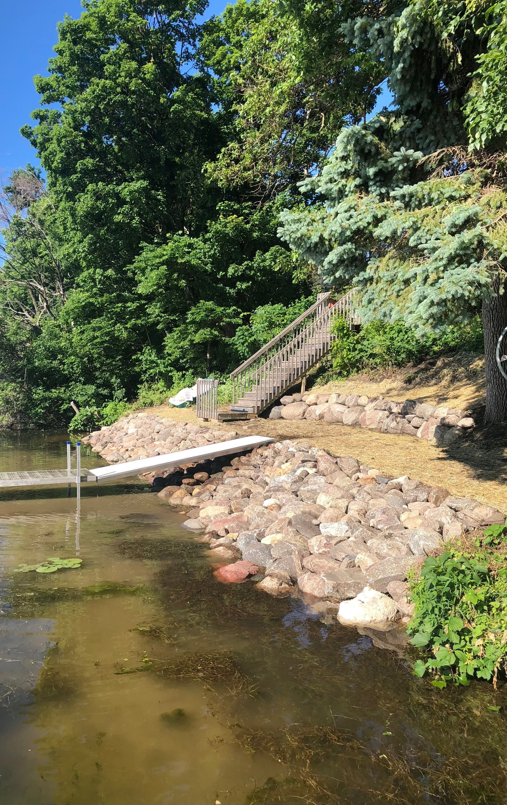 Rocky riverbank with a small dock and stairs beneath leafy green trees