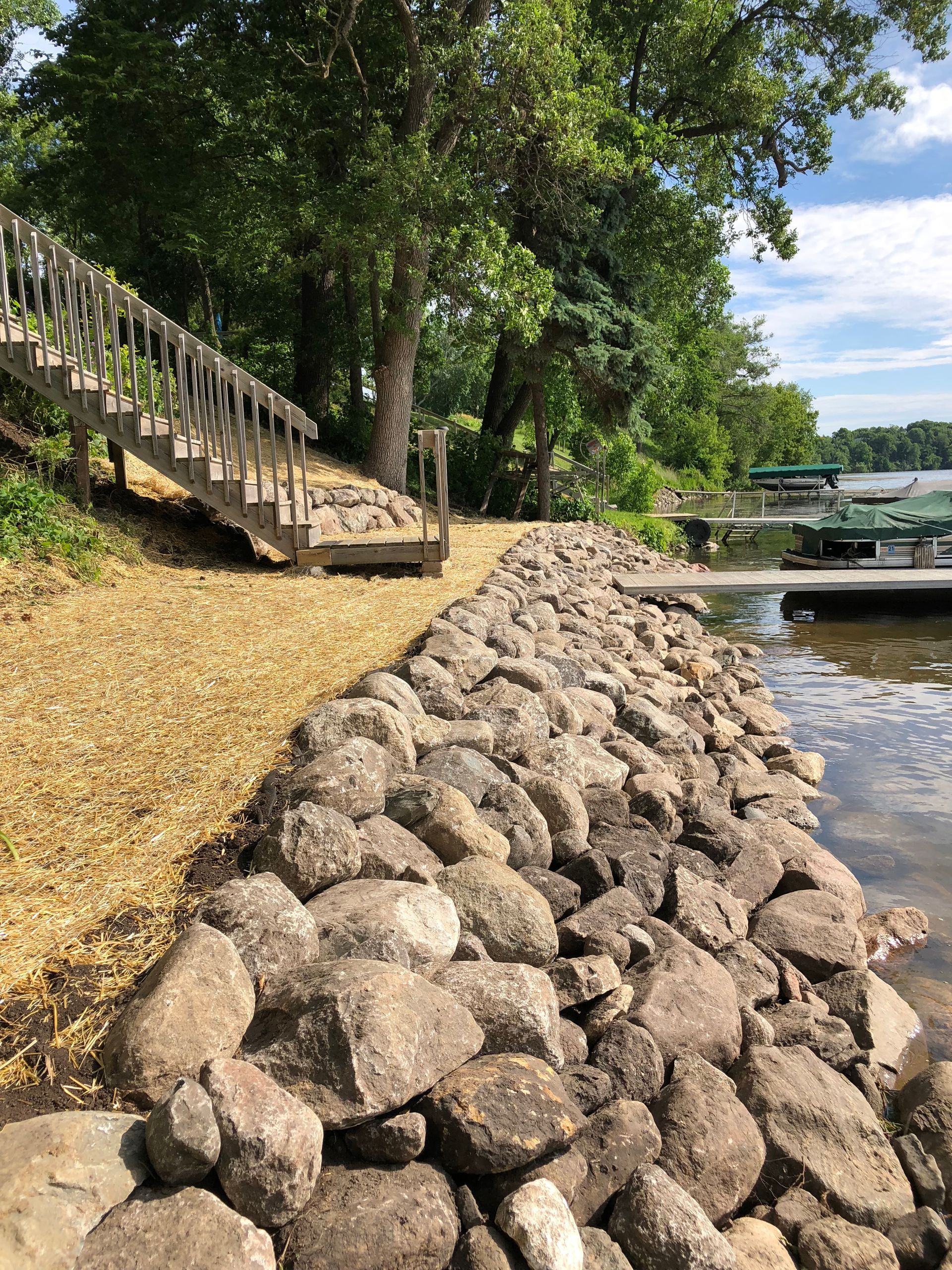 Rock-lined shoreline beside a tree-shaded path and staircase, with water and a bridge in the background