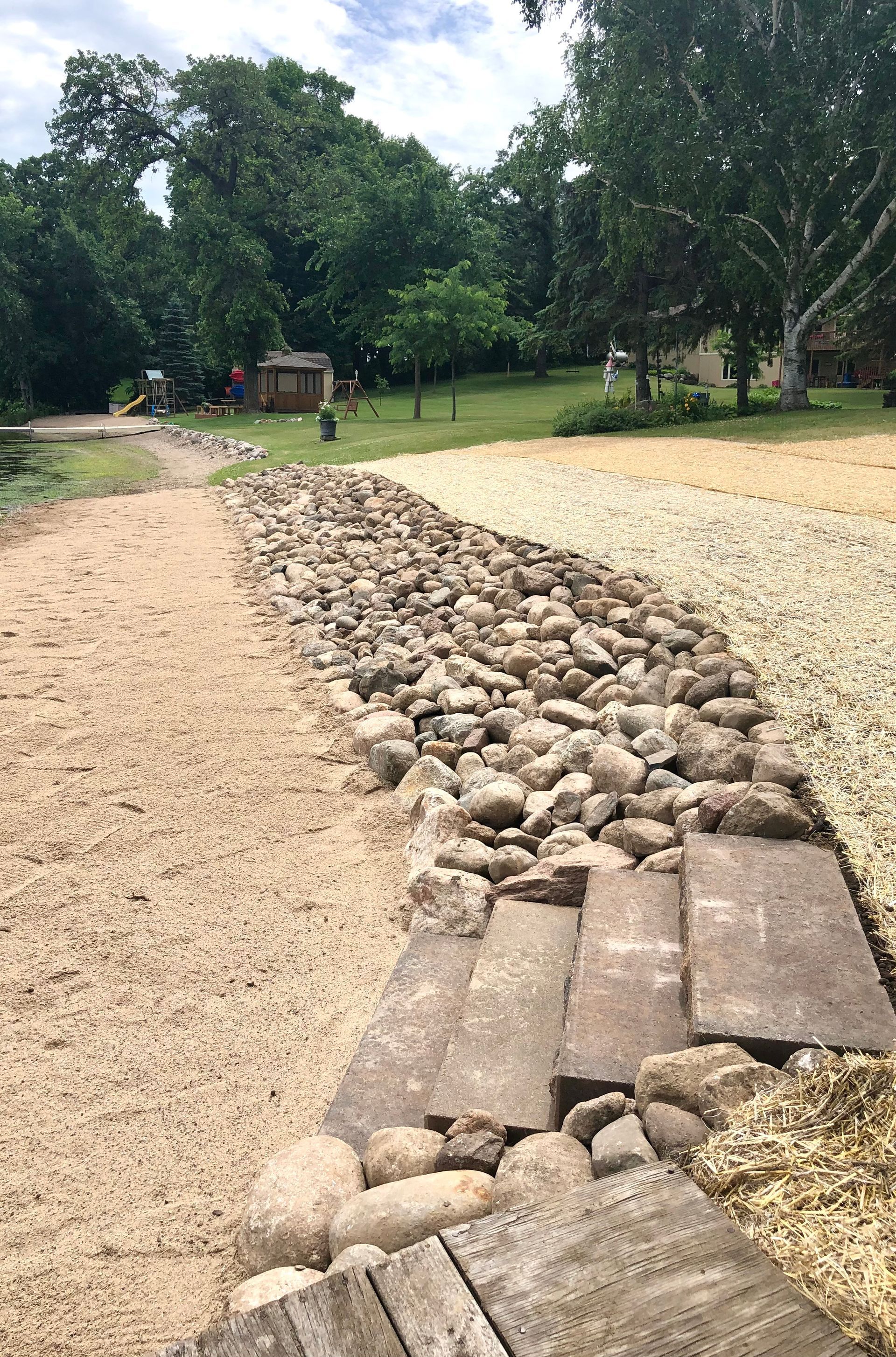 Gravel path with a stone drainage channel through a green park, with trees and a small shelter in the distance