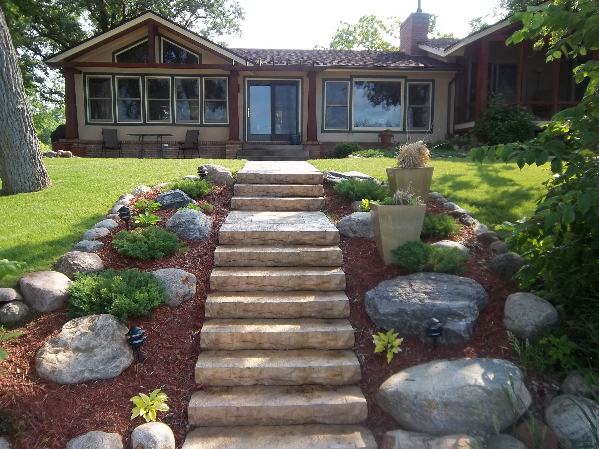 Stone steps leading up a landscaped front yard to a brown house with a porch and large windows