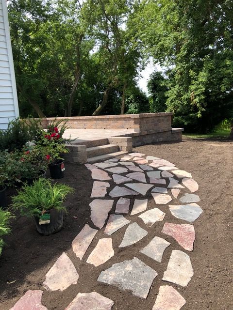 Curved stone walkway leading to a brick patio and steps beside a house and garden beds