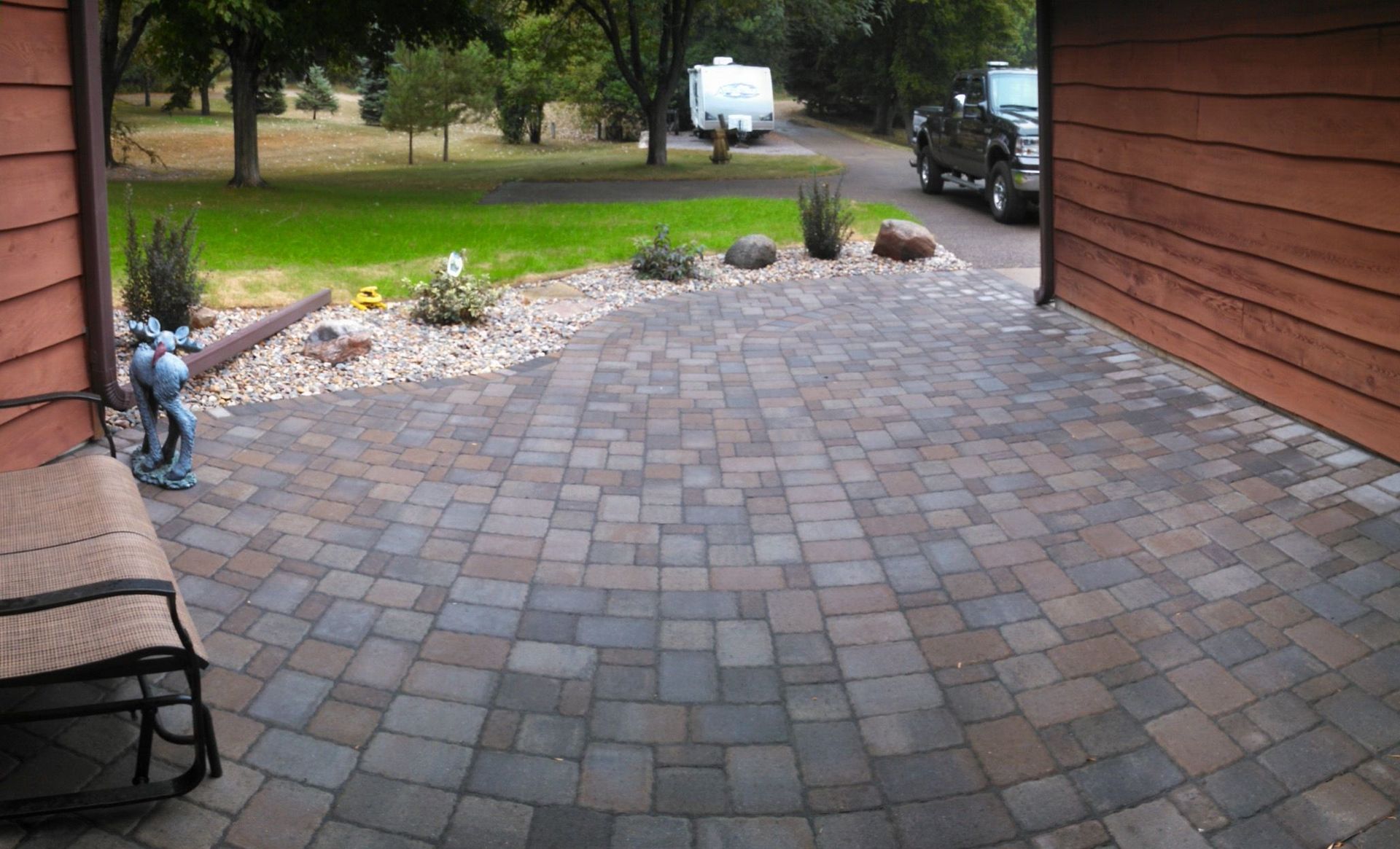 Brick patio beside a house, with a bench, bicycle, landscaping, and a driveway leading to the street.